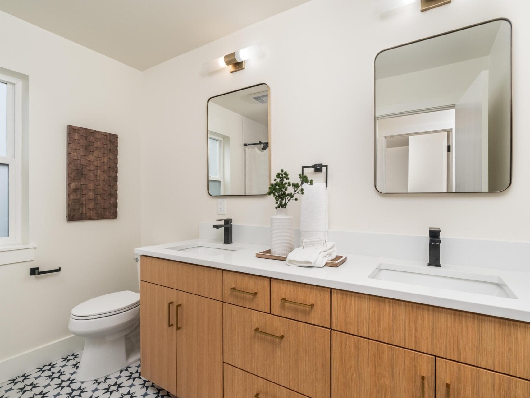 Modern bathroom with dual sinks, square mirrors, and wood cabinetry. Light fixture above each mirror. White walls, patterned black-and-white floor tiles. Toilet next to the vanity. Small plants and towels on countertop for decor.