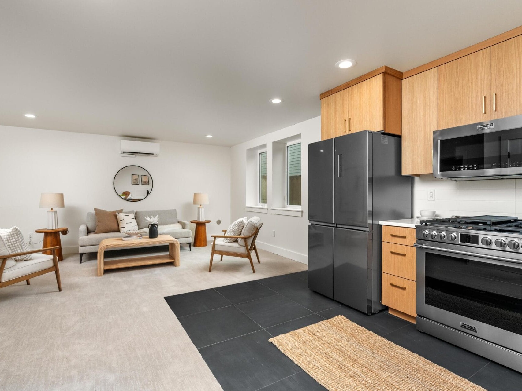 Modern open-plan living room and kitchen with light wood cabinets, stainless steel appliances, and grey tile flooring. A cozy seating area features a sofa, two chairs, and a round mirror on the wall. A woven rug separates the kitchen and living spaces.