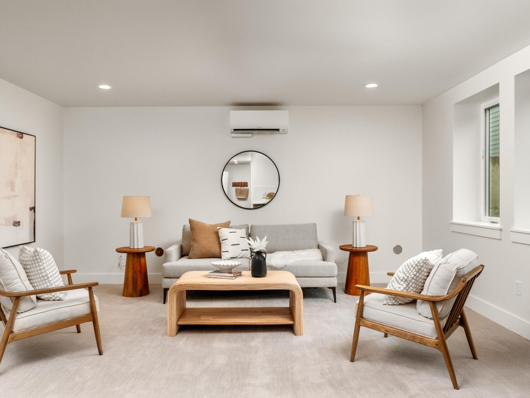 A modern living room with a gray sofa, two wooden chairs with cushions, and a wooden coffee table. A round mirror hangs above the sofa, and two lamps are on either side. Neutral tones and minimalist décor create a cozy atmosphere.