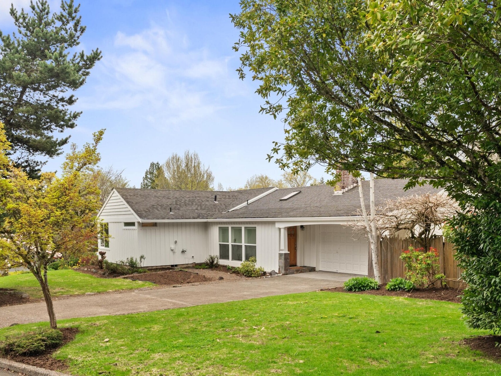 Single-story white house with a gray roof, attached garage, large front windows, and a well-kept green lawn surrounded by trees and shrubs on a clear day.
