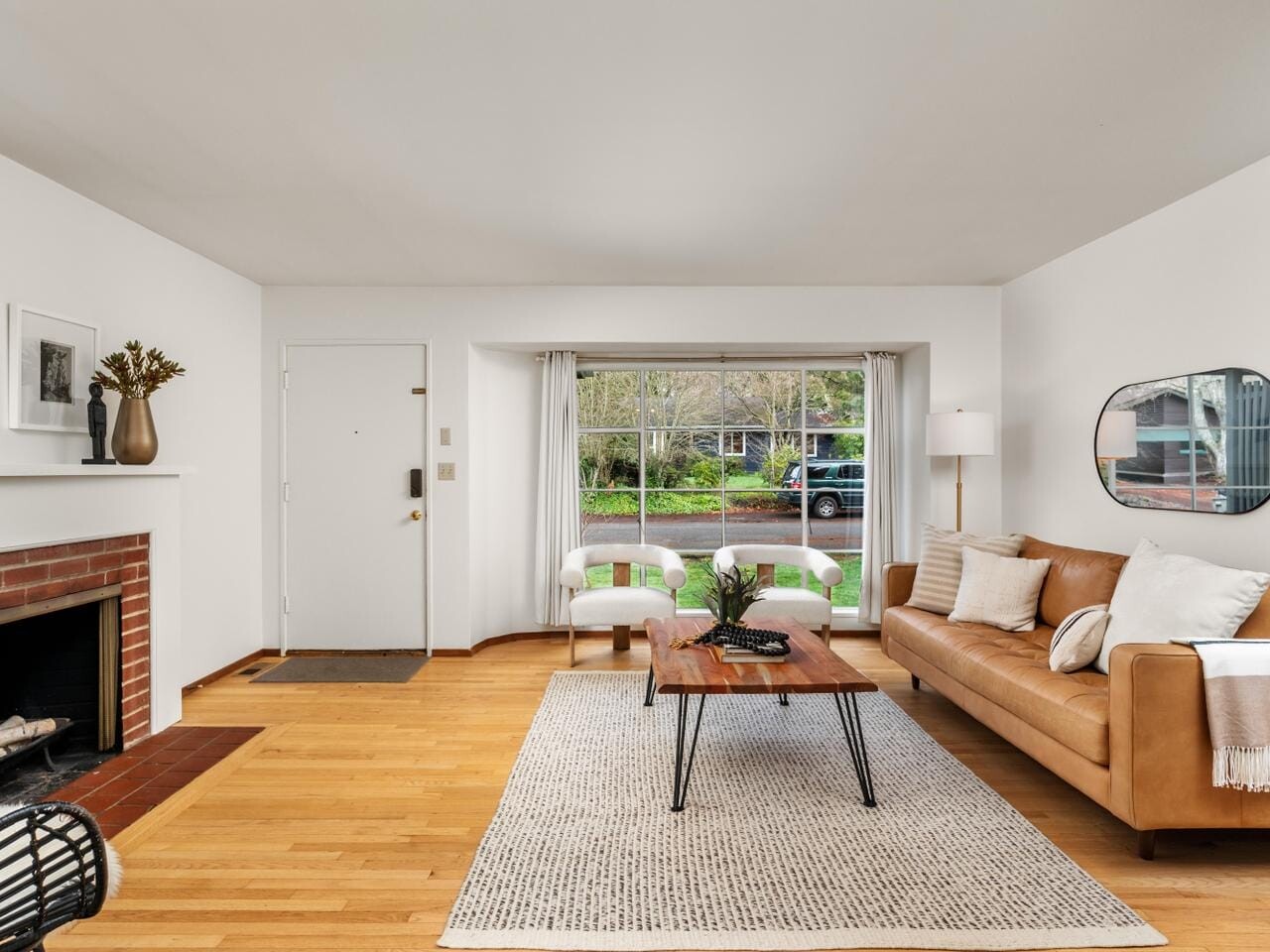 A cozy living room with a tan leather sofa, wooden coffee table, and a rug on hardwood flooring. A fireplace with a brick surround is on the left, and a large window with a view of greenery is in the background. Decor includes a mirror and wall art.