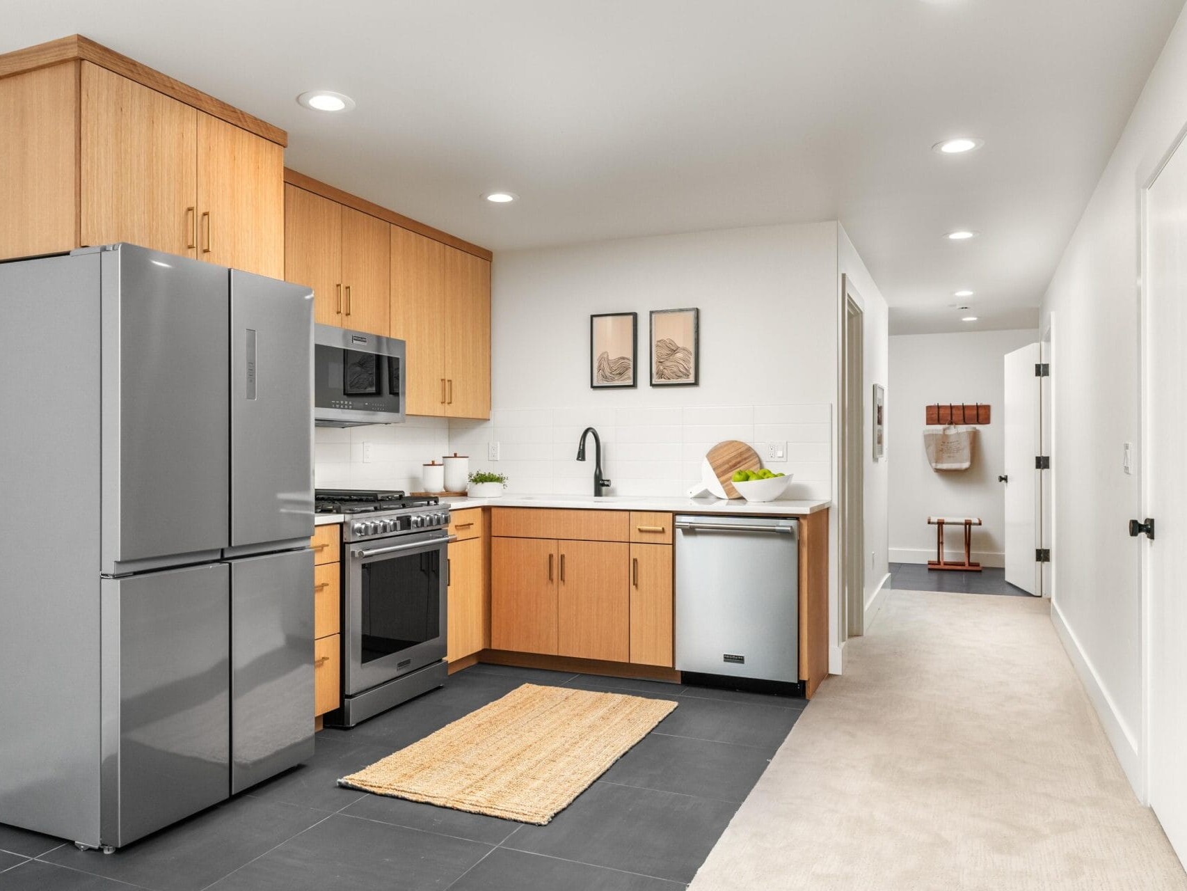 Modern kitchen with light wood cabinets, stainless steel refrigerator, oven, and dishwasher. A woven rug lies on dark tiled flooring. Two framed pictures hang on a white wall above a countertop. A hallway leads to other rooms.