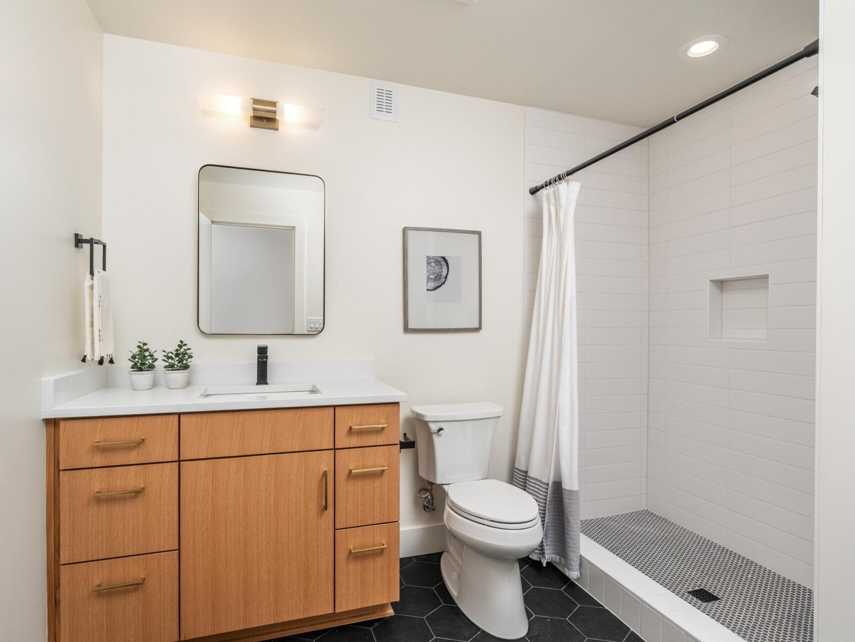 Modern bathroom with wooden vanity, black hexagonal floor tiles, and white walls. Features a wall-mounted mirror, sink with a black faucet, toilet, and a walk-in shower with a white curtain and niche. Small potted plants adorn the countertop.