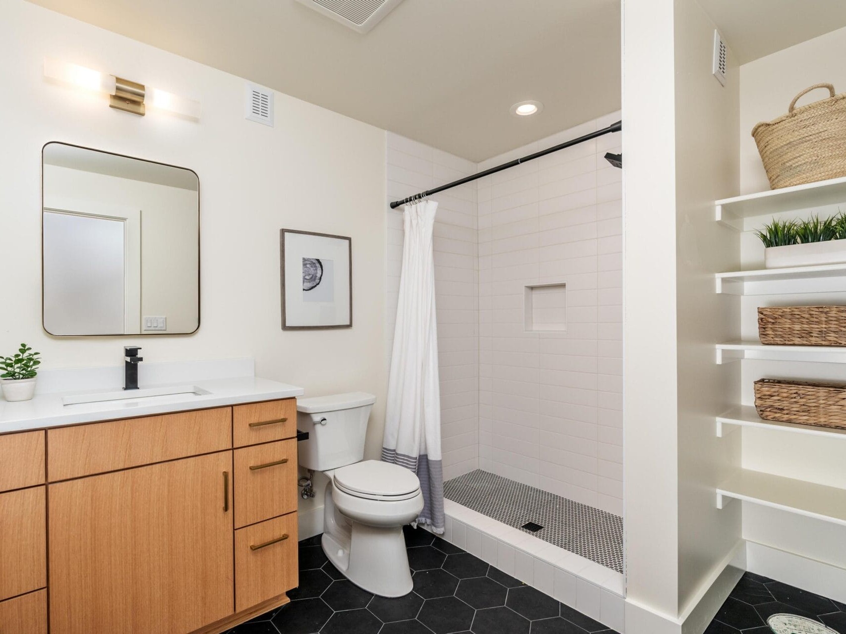 Modern bathroom with wooden vanity, white countertop, and black hexagonal floor tiles. It features a toilet, a tiled shower with a curtain, and shelves with baskets and plants. A square mirror and wall light are above the sink.