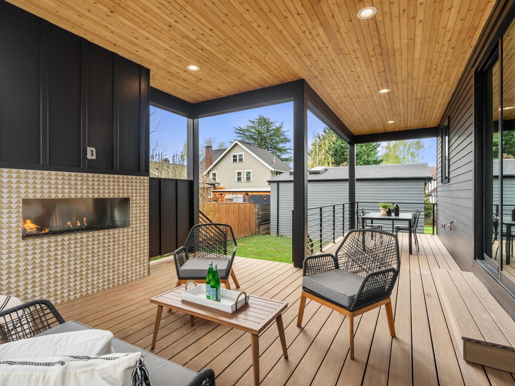 A modern covered patio with wooden ceiling and floor features a geometric-patterned fireplace, stylish wicker chairs with cushions, and a small wooden table. The patio overlooks a grassy backyard with trees and a neighboring house in the background.