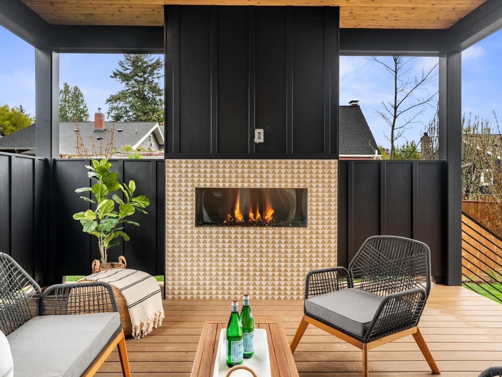 A cozy outdoor patio features a modern fireplace with a geometric tile design. Two gray cushioned chairs face the fireplace, with a small wooden table in between. A potted plant is on the left, and the area is surrounded by black railings.