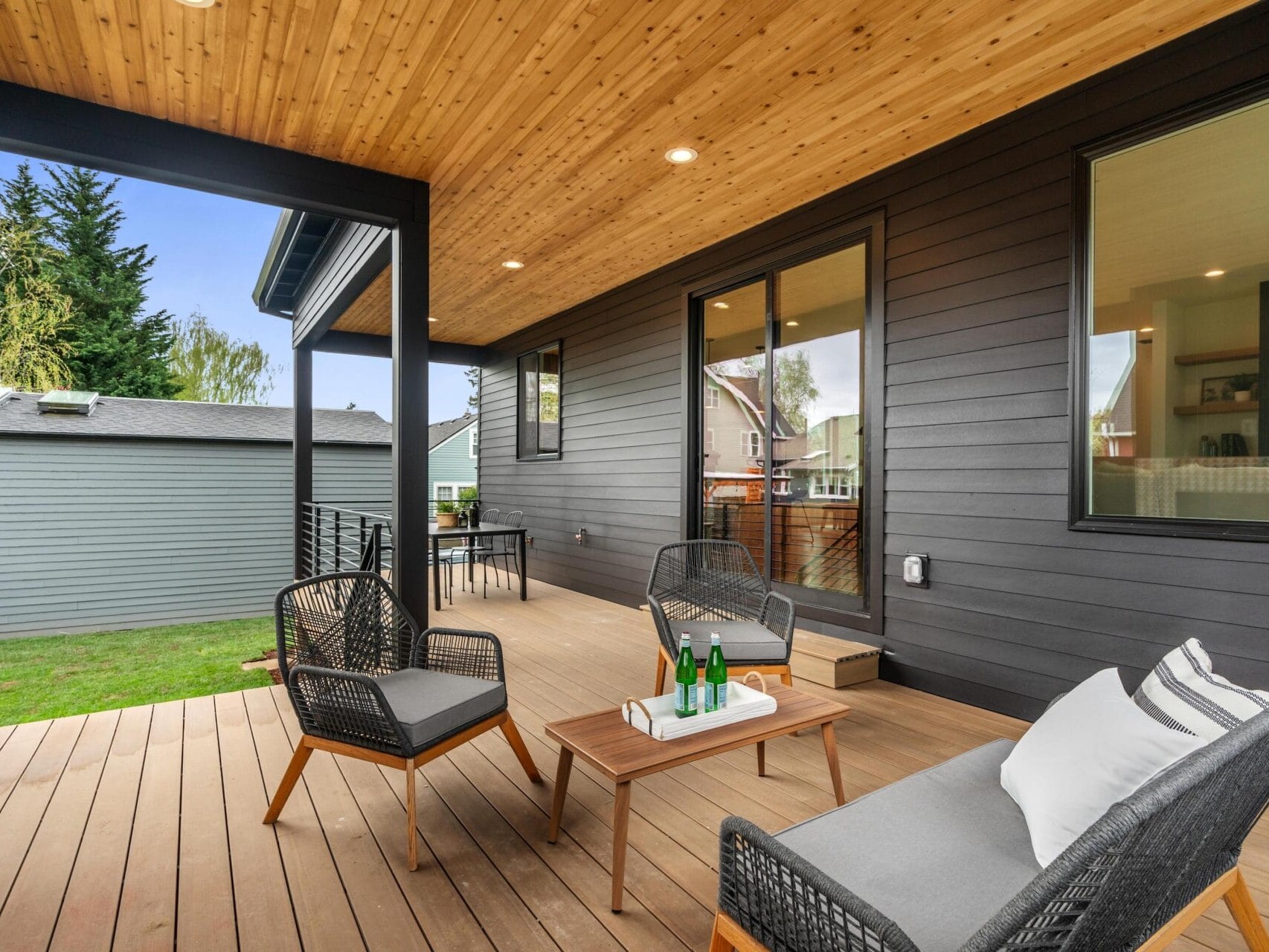 A modern wooden deck with a covered ceiling, featuring a small outdoor seating area with two chairs, a bench, and a wooden coffee table with drinks. The house has large windows, and the yard is visible in the background.