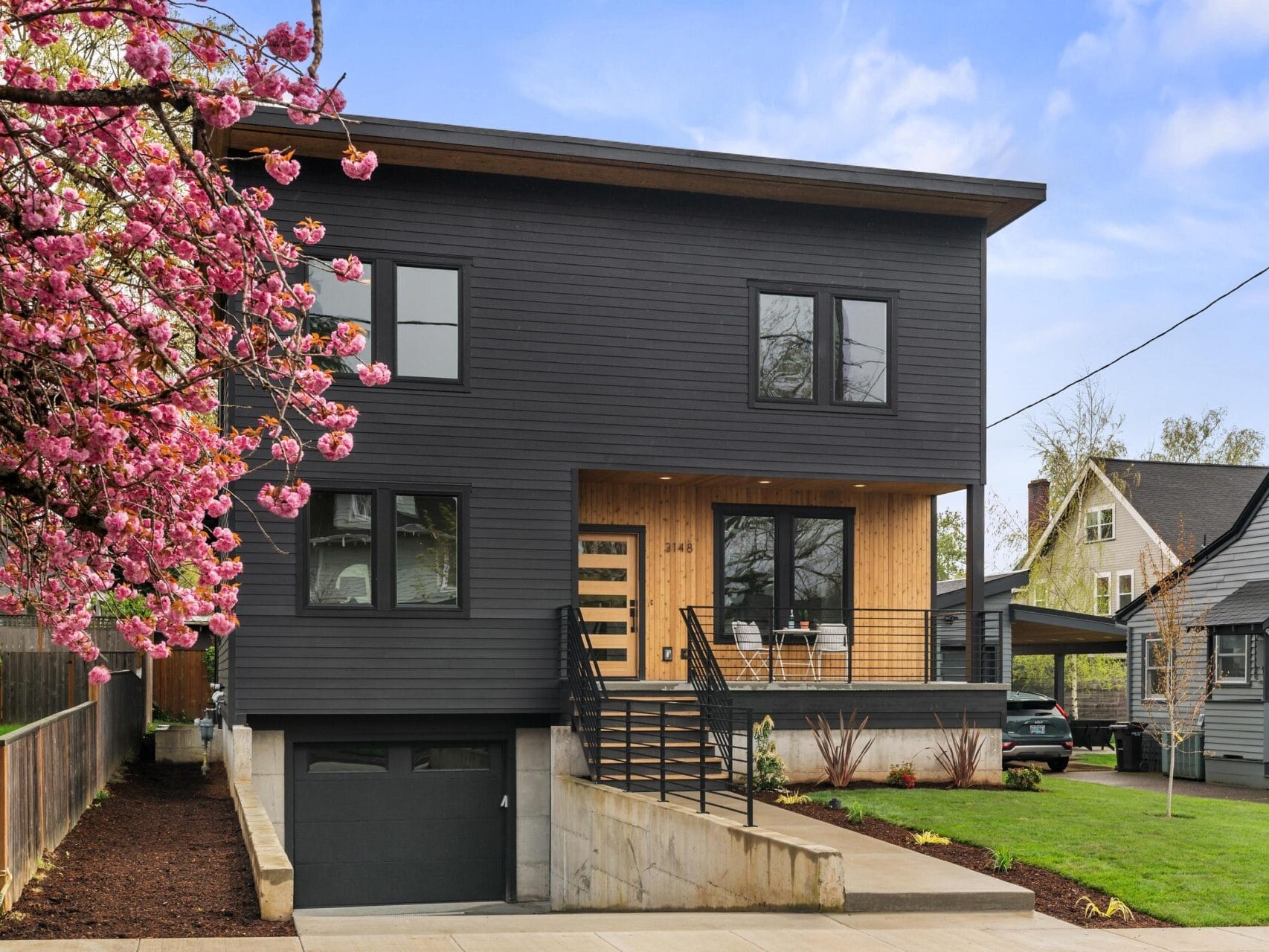 Modern two-story house with dark exterior siding, large windows, and a wooden accent around the entrance. A blossoming tree with pink flowers is on the left, and theres a short driveway leading to a garage. Neighboring homes are visible.