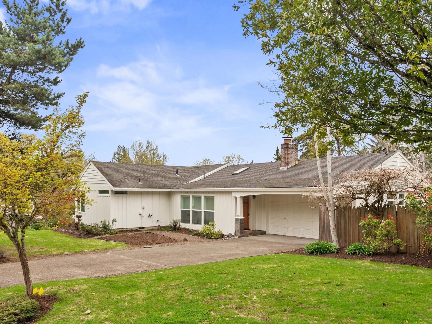 A single-story white house with a gray roof, attached garage, and large front yard with green grass and trees, under a blue sky with scattered clouds.