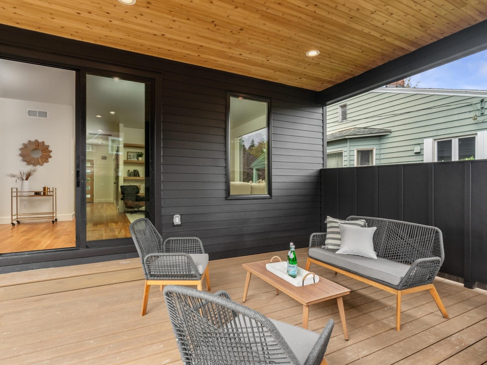 Covered wooden deck with two modern gray chairs, a matching loveseat, and a wooden coffee table. A bottle and two glasses are on the table. The area is enclosed by a black fence and theres a large sliding door leading into a house.