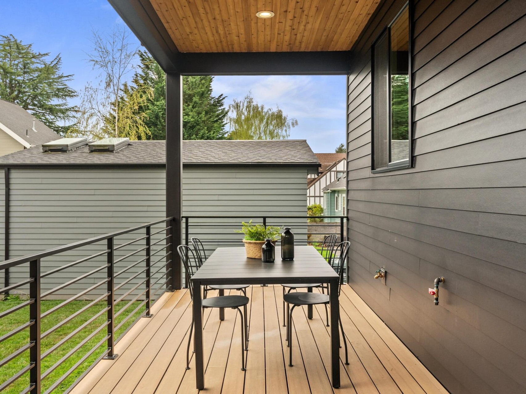 A modern outdoor patio with a wooden deck features a rectangular black table and four matching chairs. Potted plants sit on the table. The surrounding area has a view of trees and neighboring houses under a blue sky.