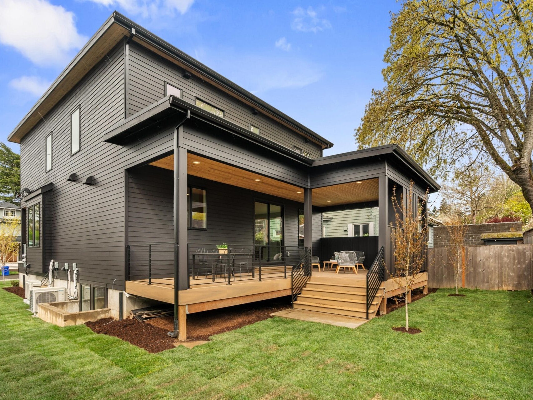 A modern two-story house with dark wood siding and a spacious, elevated wooden deck. The deck features patio furniture and is surrounded by a well-manicured lawn and trees. The sky is clear and blue.