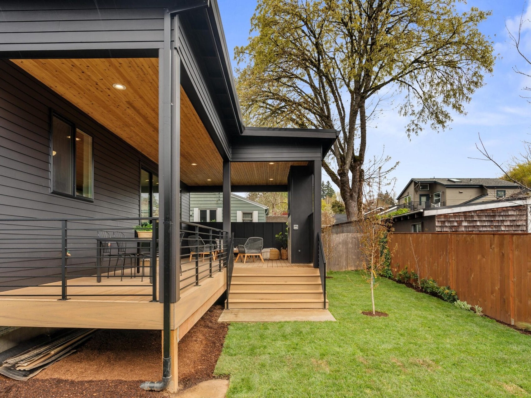 A modern house with a covered wooden patio featuring outdoor seating and dining furniture. The lawn is neatly maintained, and a wooden fence separates the yard from neighboring houses. A large tree is visible near the fence.