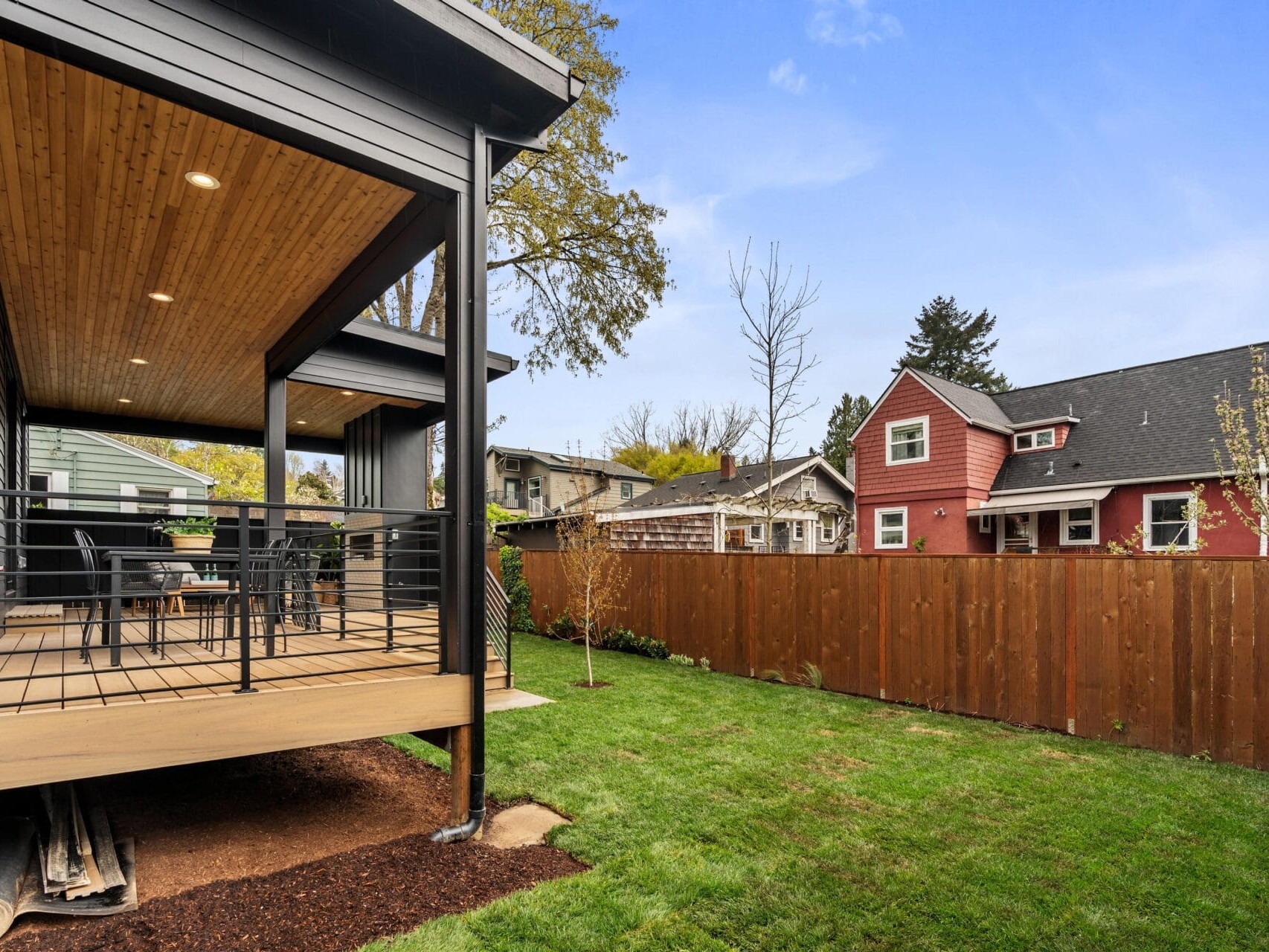 A backyard with a grass lawn and a wooden fence. To the left, theres a covered wooden deck with patio furniture and black railing attached to a modern house. In the background, other houses are visible under a clear blue sky.