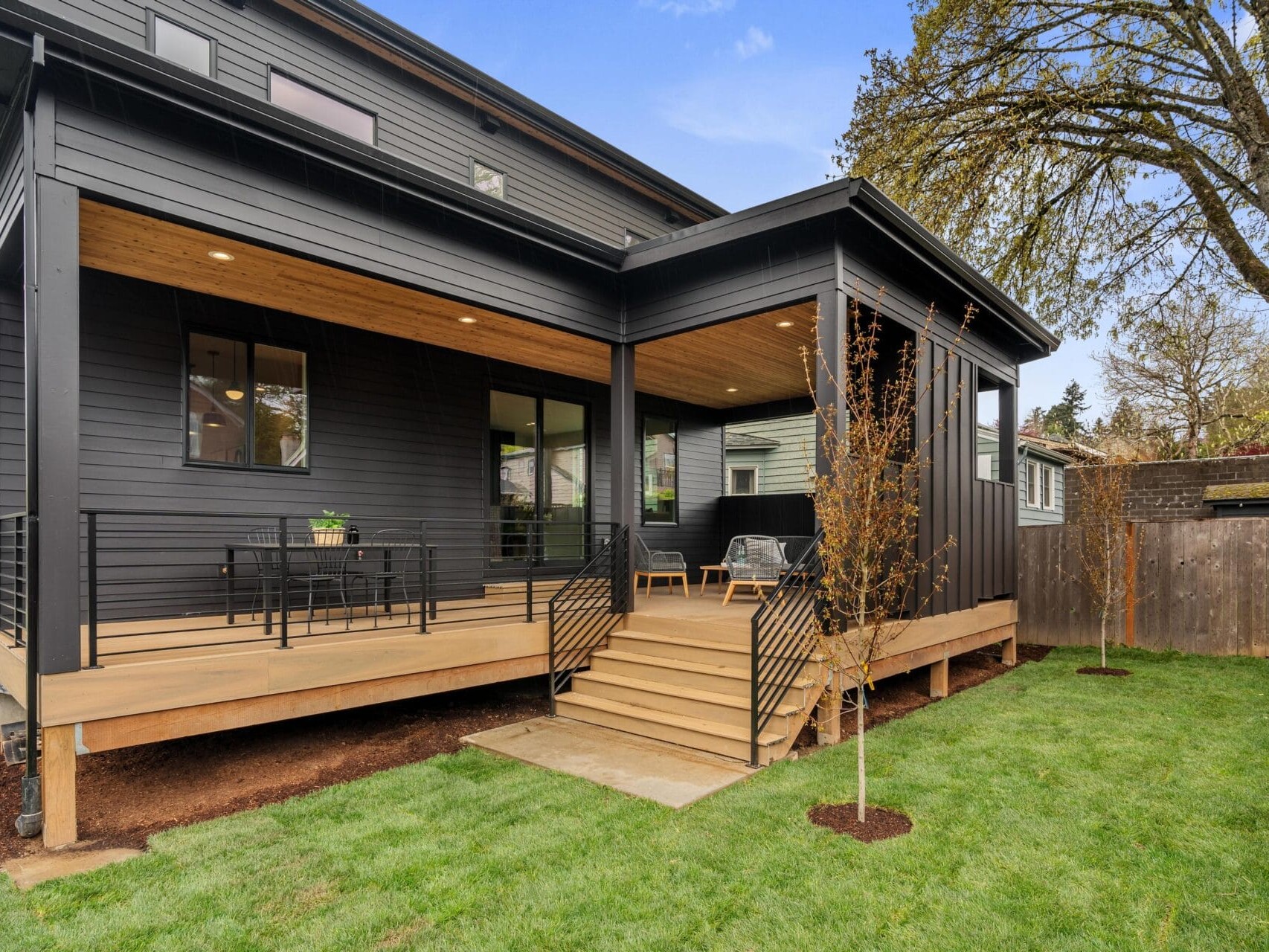 Modern black house with a spacious wooden deck featuring outdoor seating and a table with chairs. The deck is surrounded by a neatly manicured lawn and a few young trees, with a fenced backyard under a clear blue sky.