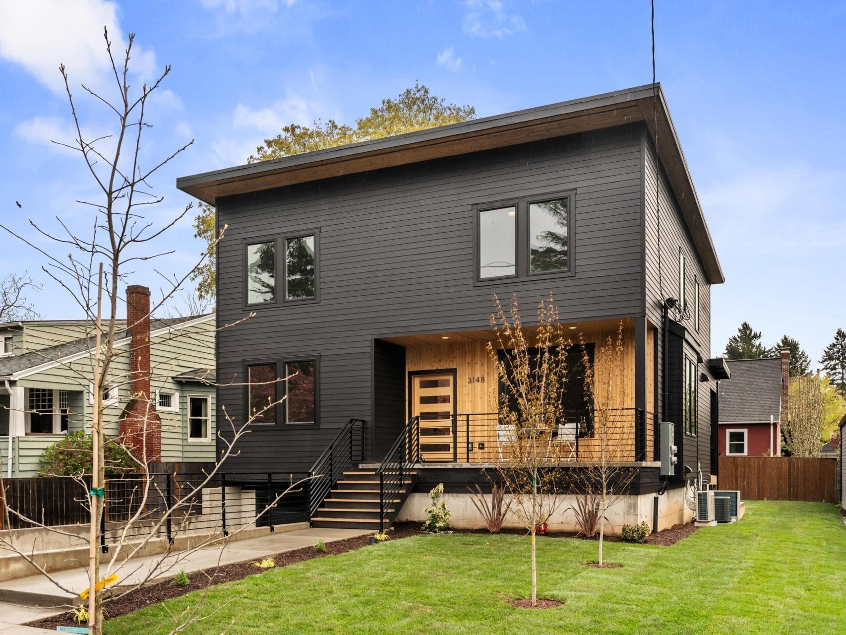 Modern two-story house with dark gray siding and wooden accents. Features multiple large windows and a welcoming porch. Surrounded by a well-maintained lawn and young trees, set in a residential neighborhood under a clear blue sky.