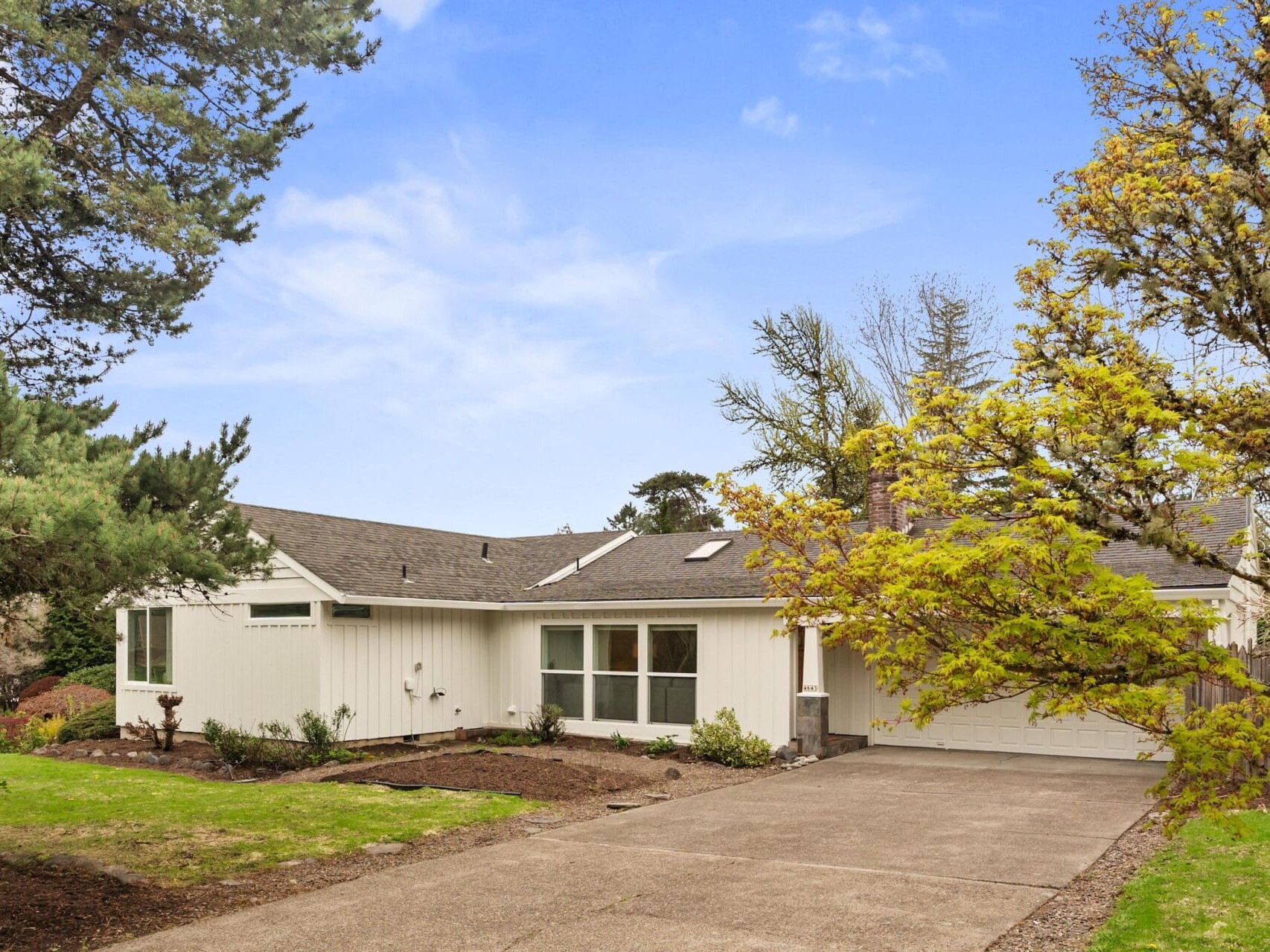 Single-story white house with a gray roof, large front windows, and an attached garage, surrounded by trees and a spacious driveway under a clear blue sky.