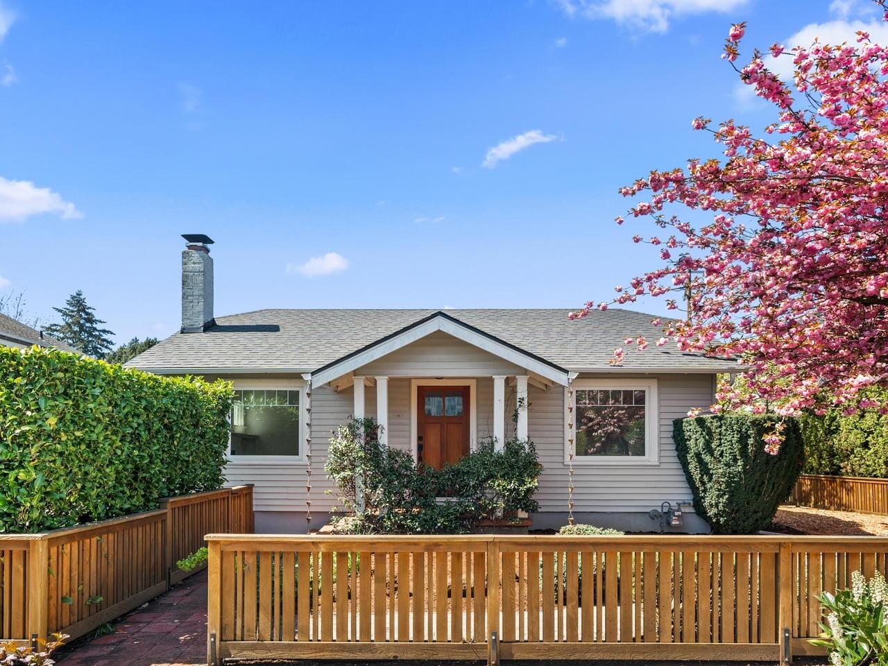 Charming single-story house with beige siding, a front porch, and a wooden fence. A blooming pink tree is on the right, lush greenery surrounds the yard, and the sky is blue with a few clouds.