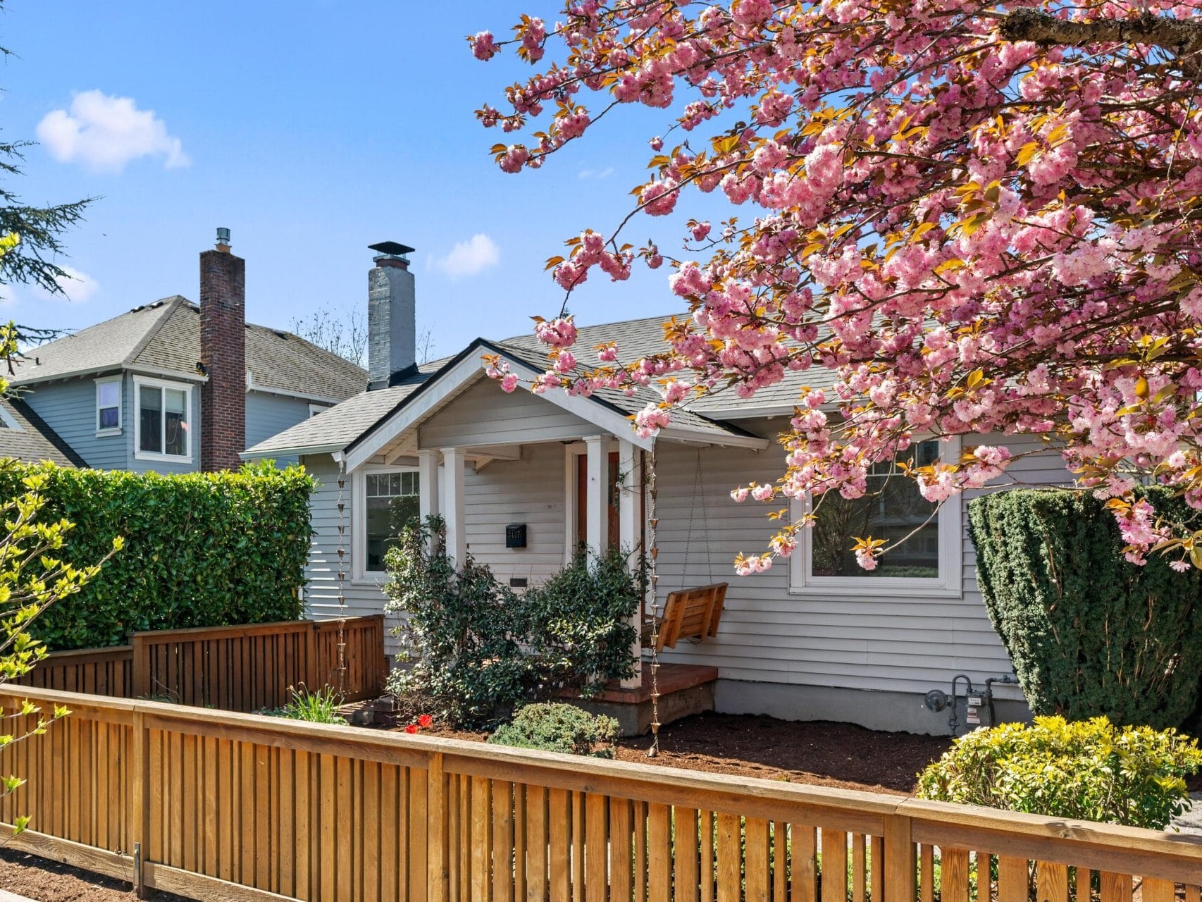 A small, light gray house with a front porch and a wooden fence, surrounded by blooming pink cherry blossom trees and green shrubs, on a sunny day.