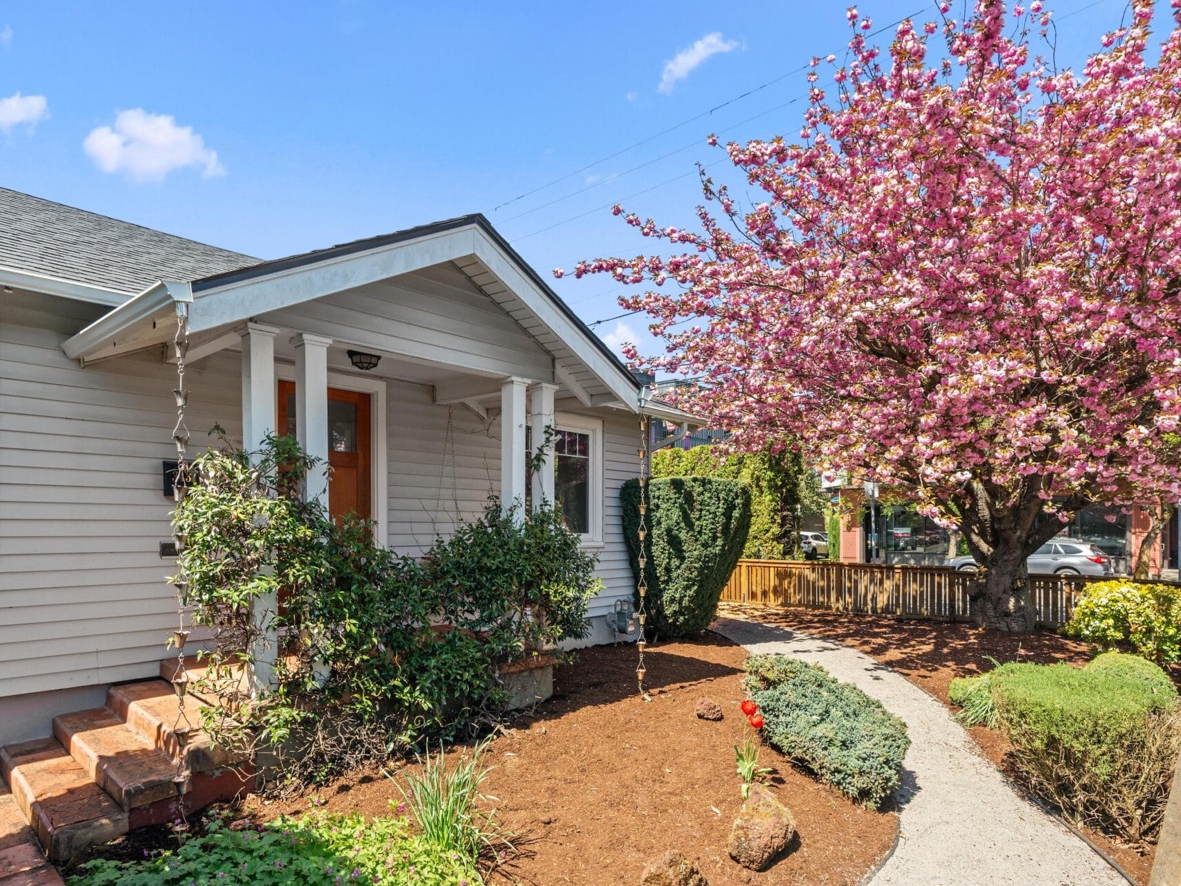Single-story house with light siding and a small front porch, surrounded by a landscaped yard. A path leads to the door, and a large tree with pink blossoms is in full bloom beside the house.