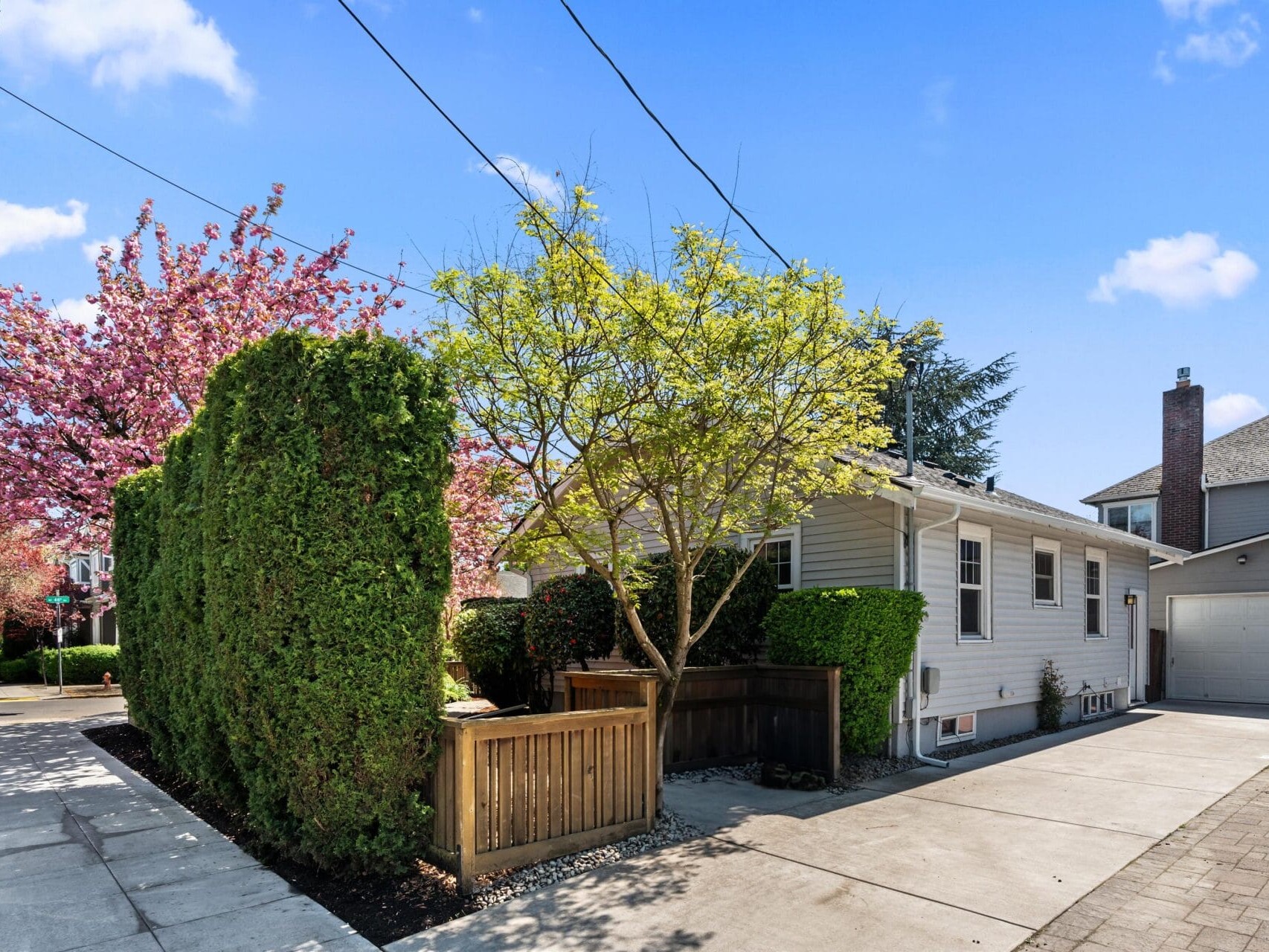 A small gray house with white trim sits on a sunny corner lot, surrounded by blooming trees and greenery. A wooden fence and neatly trimmed bushes line the driveway, with a detached garage visible in the background.