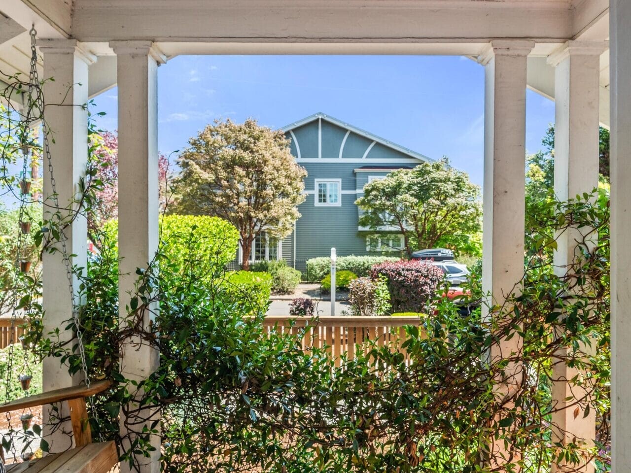View from a porch looking out at a garden and a green house across the street, with white columns, leafy vines, a wooden bench, and a tiled floor under a black porch light.