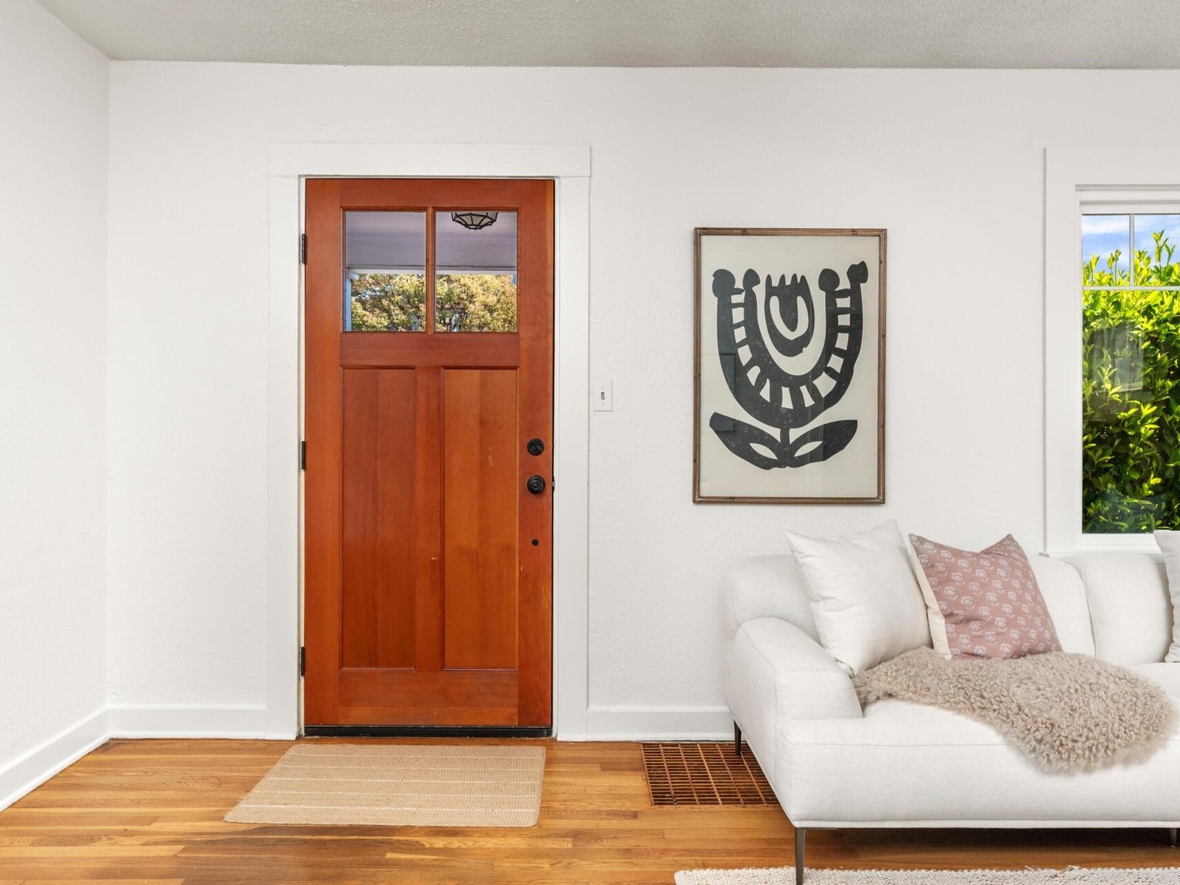 A bright entryway with a wooden front door, a beige rug, a white couch with pink and beige pillows and a throw, modern wall art, and a window showing greenery outside.