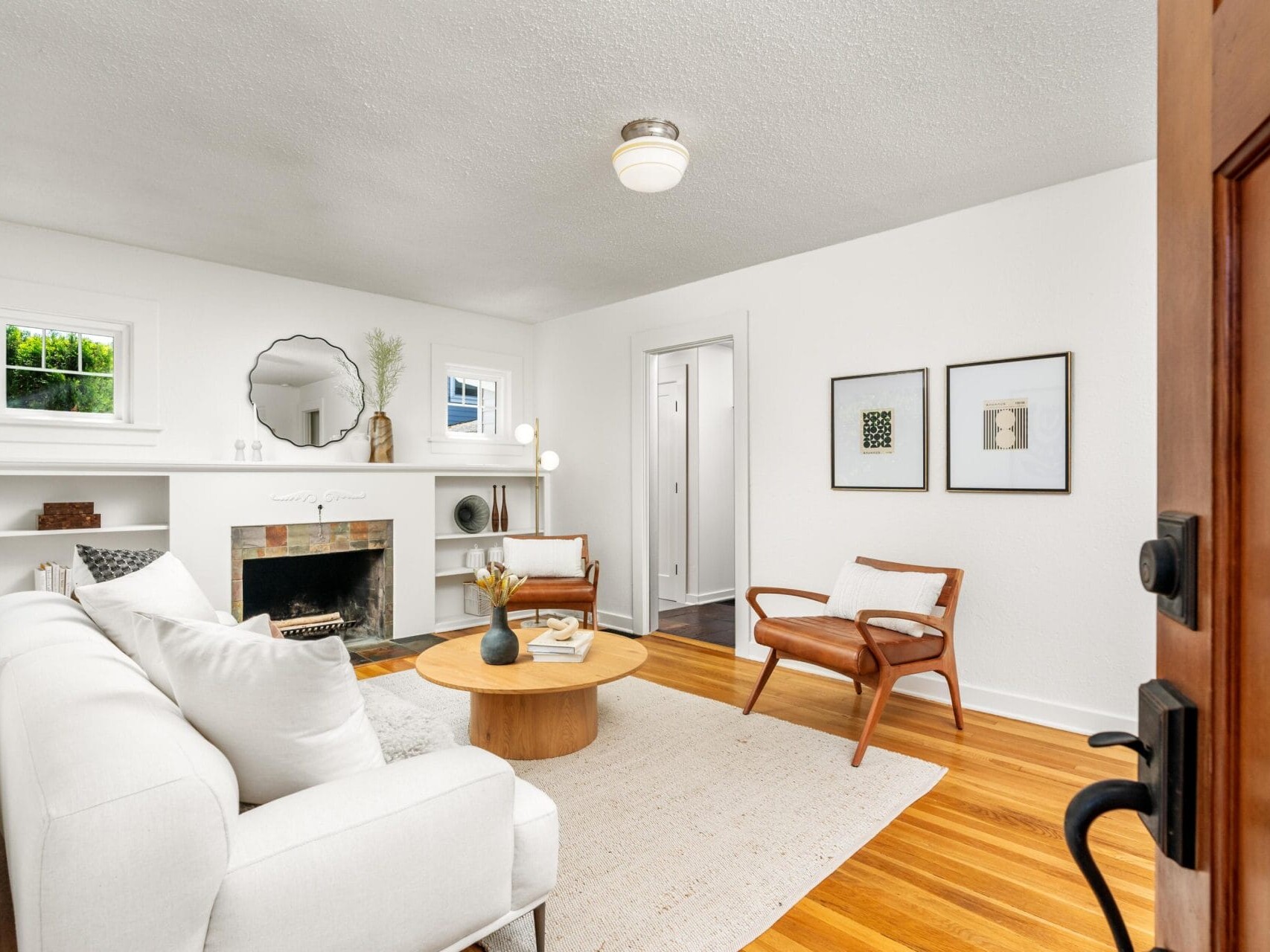 A bright living room with white walls, a white sofa, a round wooden coffee table, two wooden chairs, a fireplace, built-in shelves, framed art on the wall, and a light wood floor.