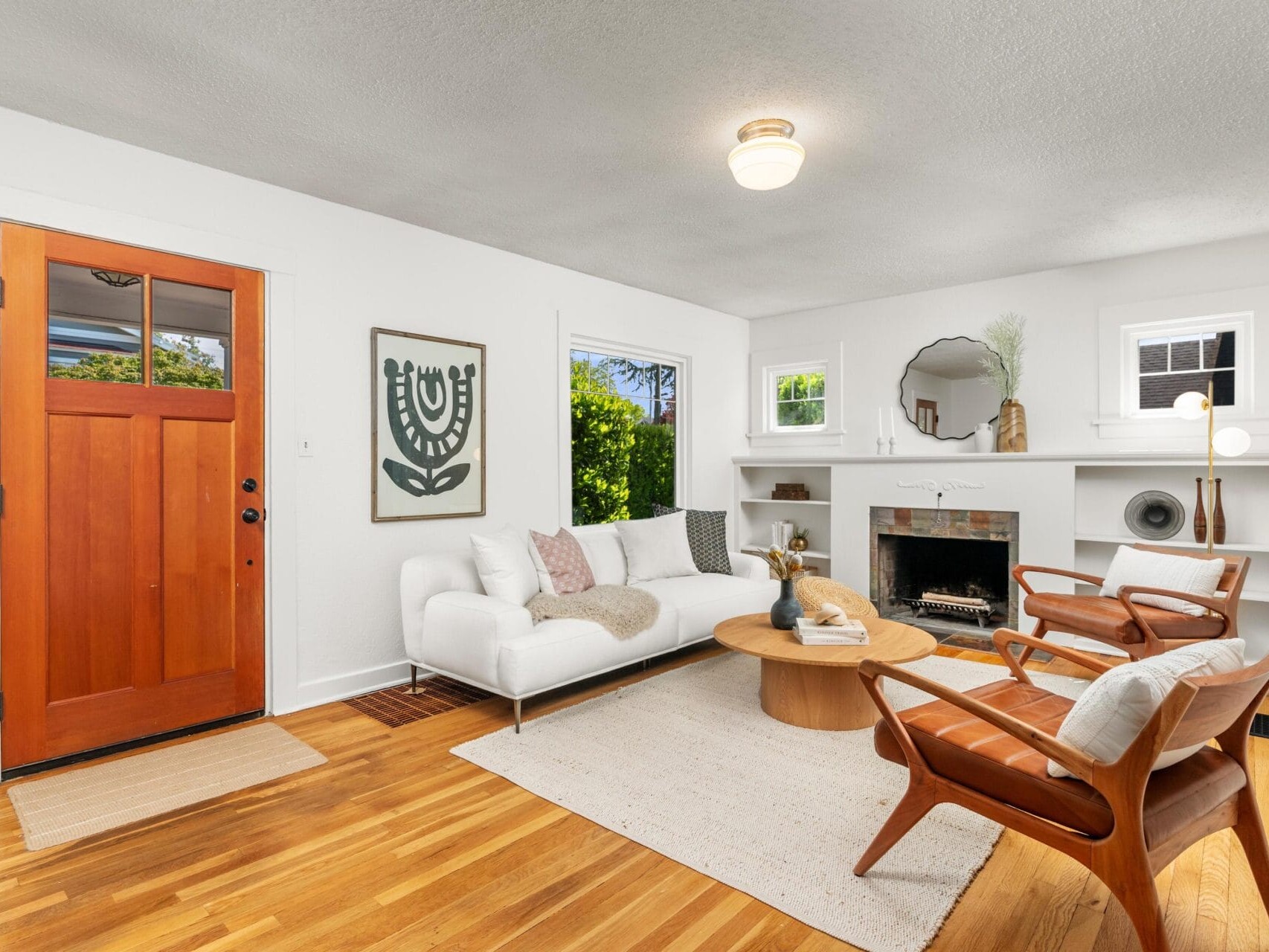 Bright, modern living room with a wooden front door, white walls, light wood floors, a white sofa, two wooden chairs, a round coffee table, a fireplace, and minimalist decor. Large windows let in natural light.