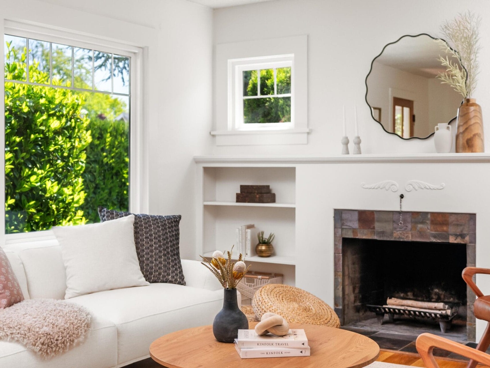 Bright living room with a white sofa, throw pillows, a round wooden coffee table, decorative vases, a fireplace with a mirror above, built-in shelves, and large windows letting in natural light.