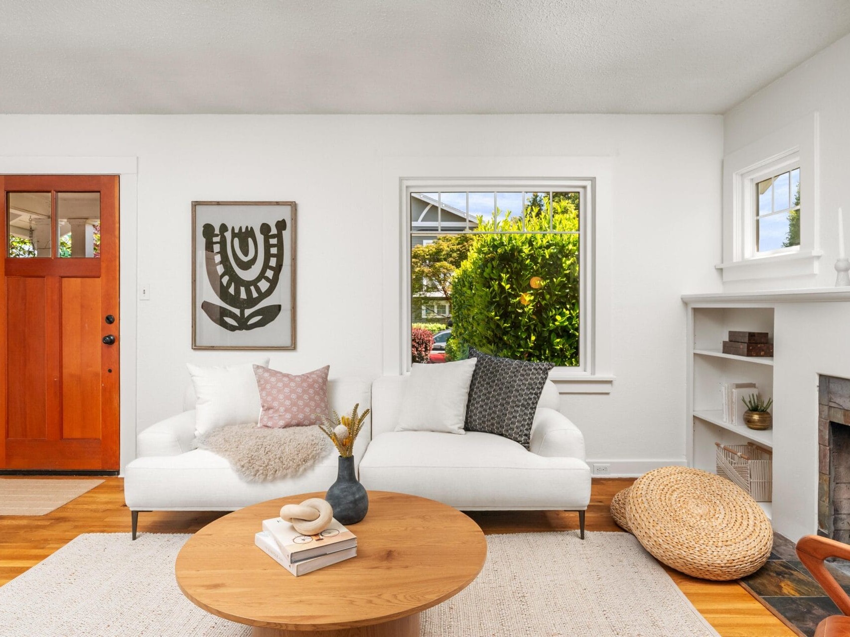 A bright living room with a white sofa adorned with pillows and a throw, a round wooden coffee table, a fireplace with built-in shelves, a large window, and a wooden front door.