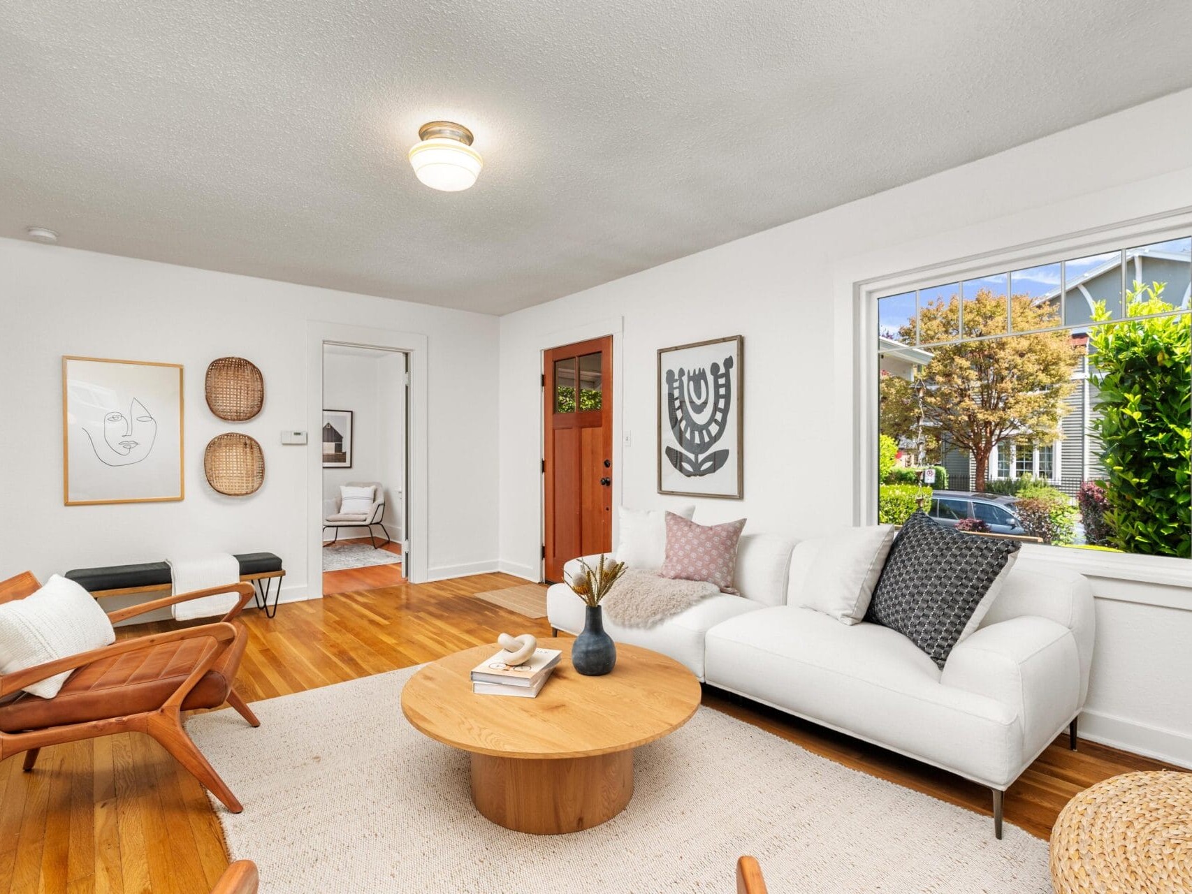Bright living room with a white sofa, wooden chairs, round coffee table, and neutral decor. Large window brings in natural light and shows greenery outside. Hardwood floors and modern, minimalist style throughout.