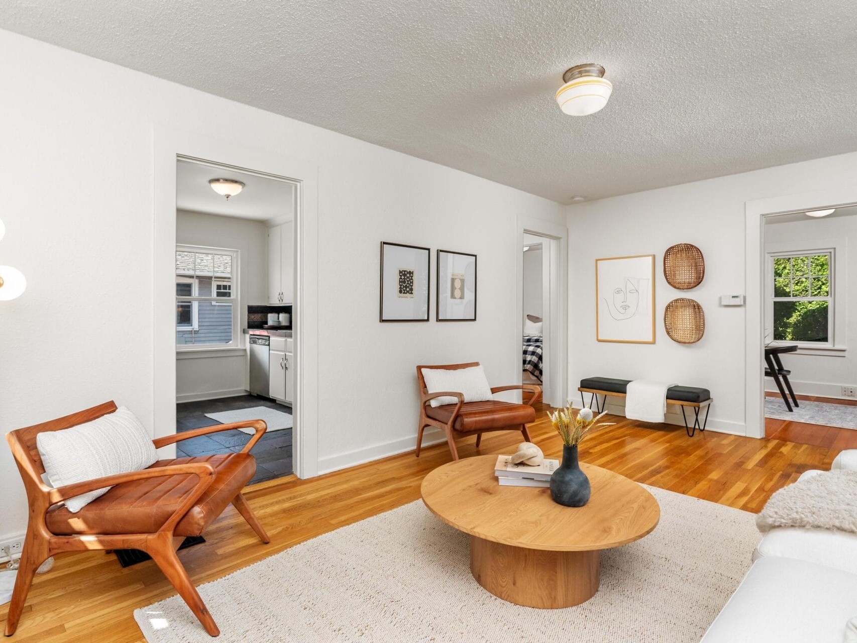 A bright living room with wooden flooring, a round wooden coffee table, two modern wooden chairs with white cushions, a white sofa, and minimal wall art. Doorways lead to a kitchen and other rooms.