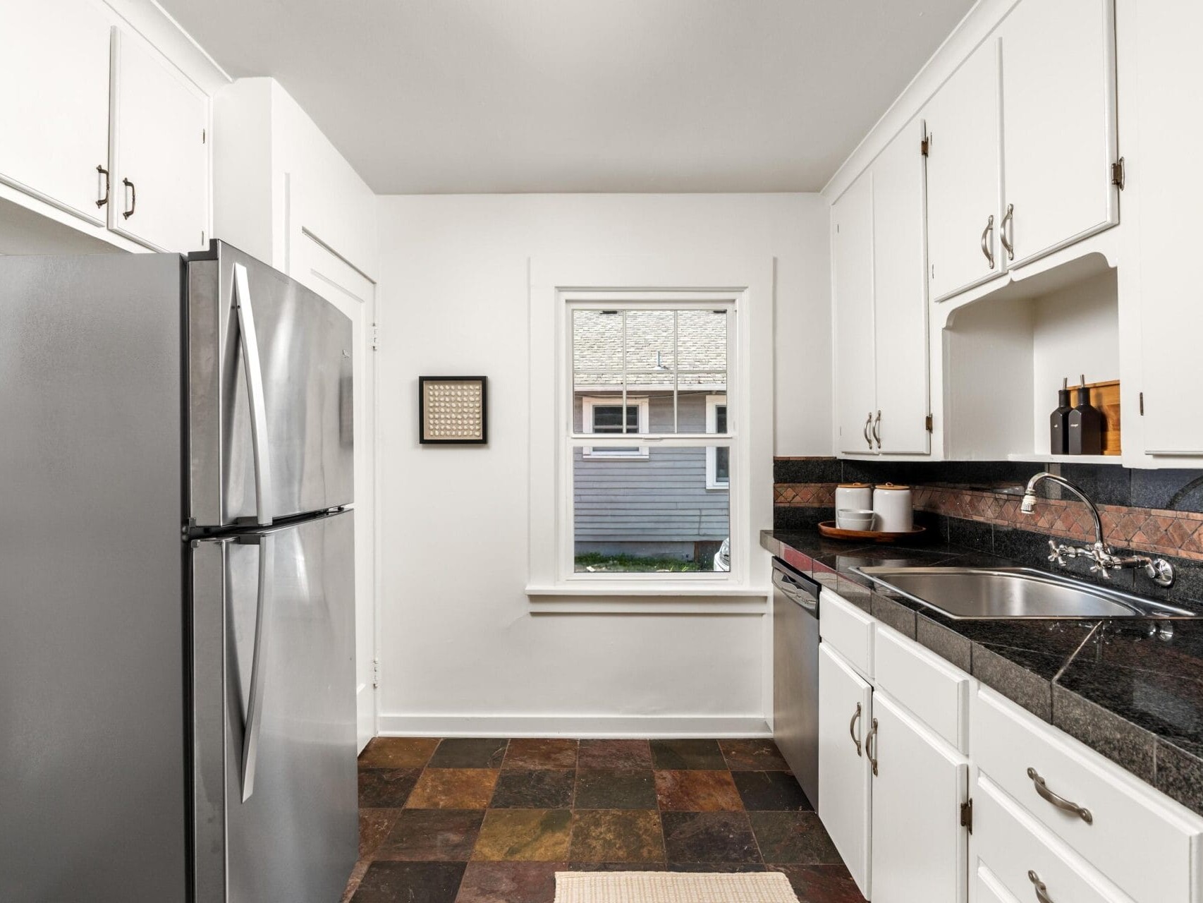 A kitchen with white cabinets, stainless steel refrigerator, dark countertops, a sink, and a window overlooking a house. The floor is tiled in earth tones, and kitchen items are neatly arranged.