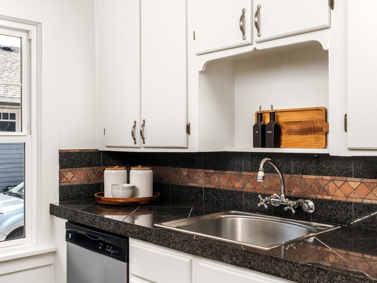 A kitchen with white cabinets, a stainless steel dishwasher, black countertops, a double sink, and a backsplash with brown and black tile accents. A cutting board and spice jars sit near the sink.