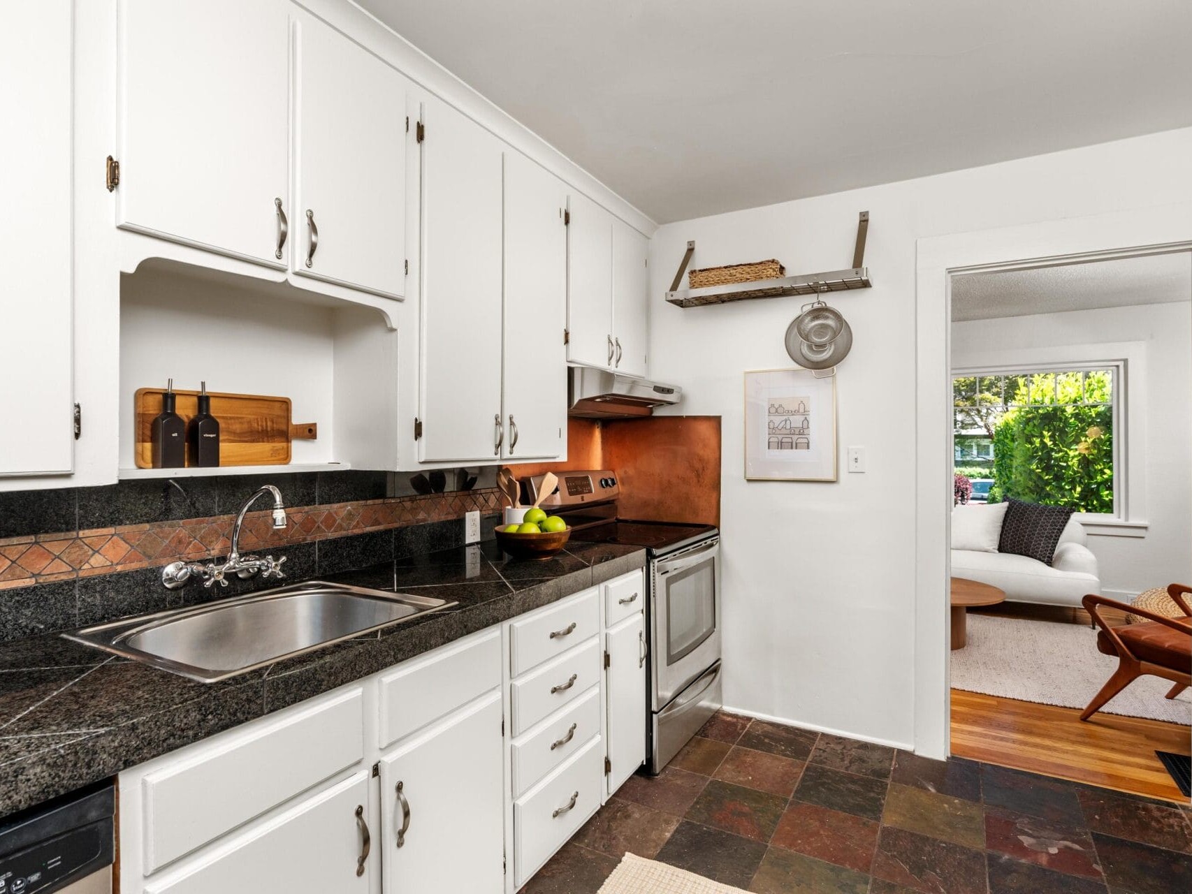 A galley-style kitchen with white cabinets, dark granite countertops, stainless steel appliances, and a tiled floor. The kitchen opens into a bright living room with a couch by a large window.
