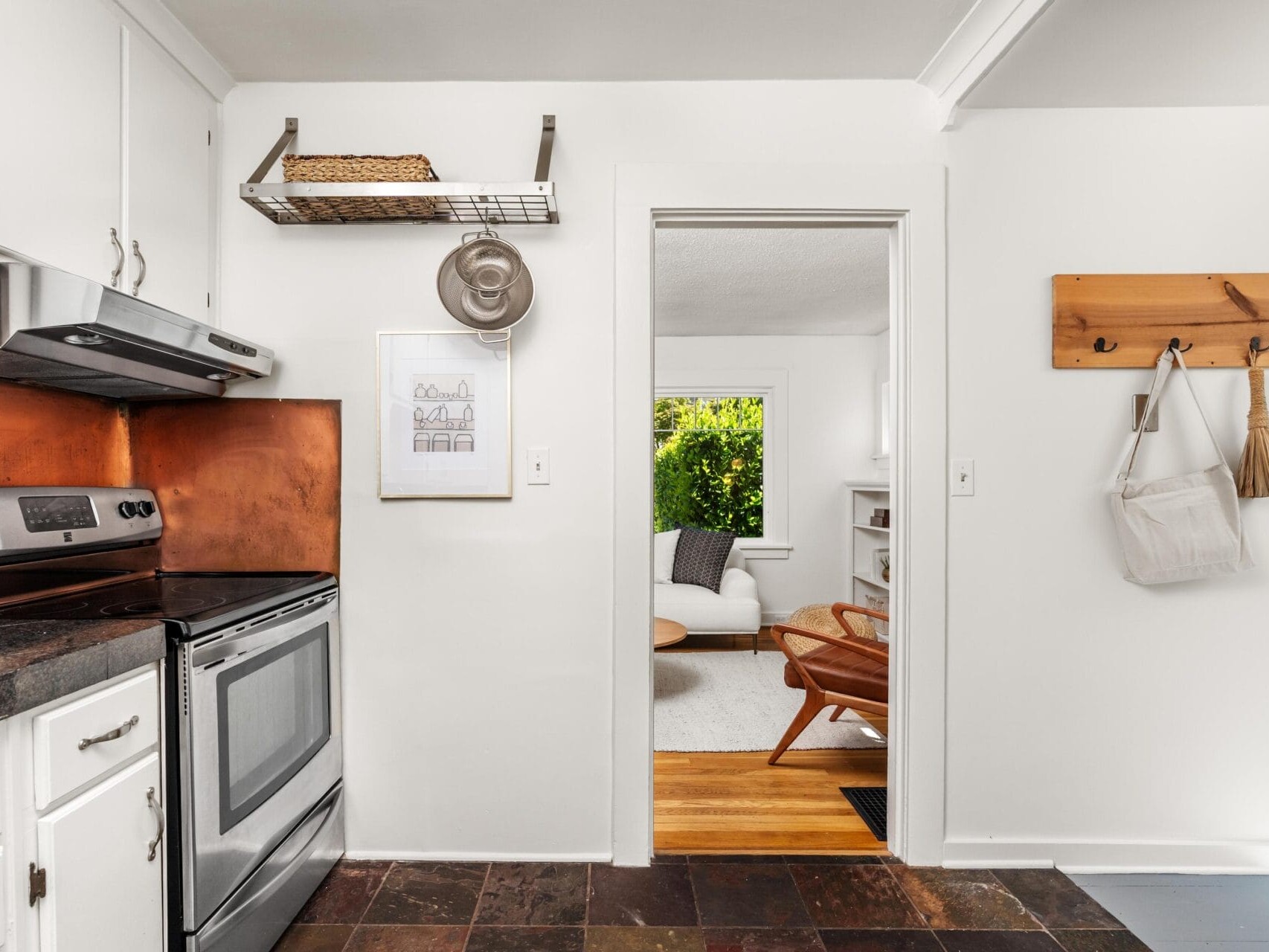 A bright kitchen with white cabinets, a stainless steel stove, a copper backsplash, and dark tile floors opens into a cozy living room with a wood chair, white sofa, and large window with greenery outside.