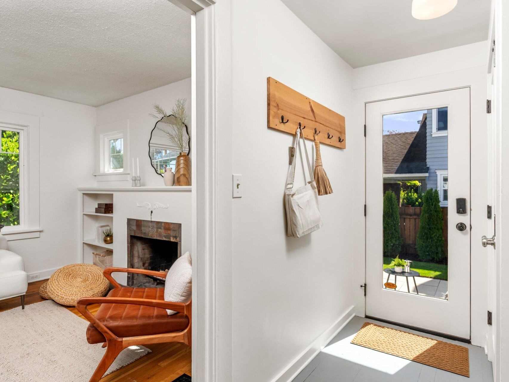 A bright entryway with white walls, a wooden coat rack holding bags, and a glass door leading outside; to the left is a cozy living room with a fireplace, armchair, and woven decor.