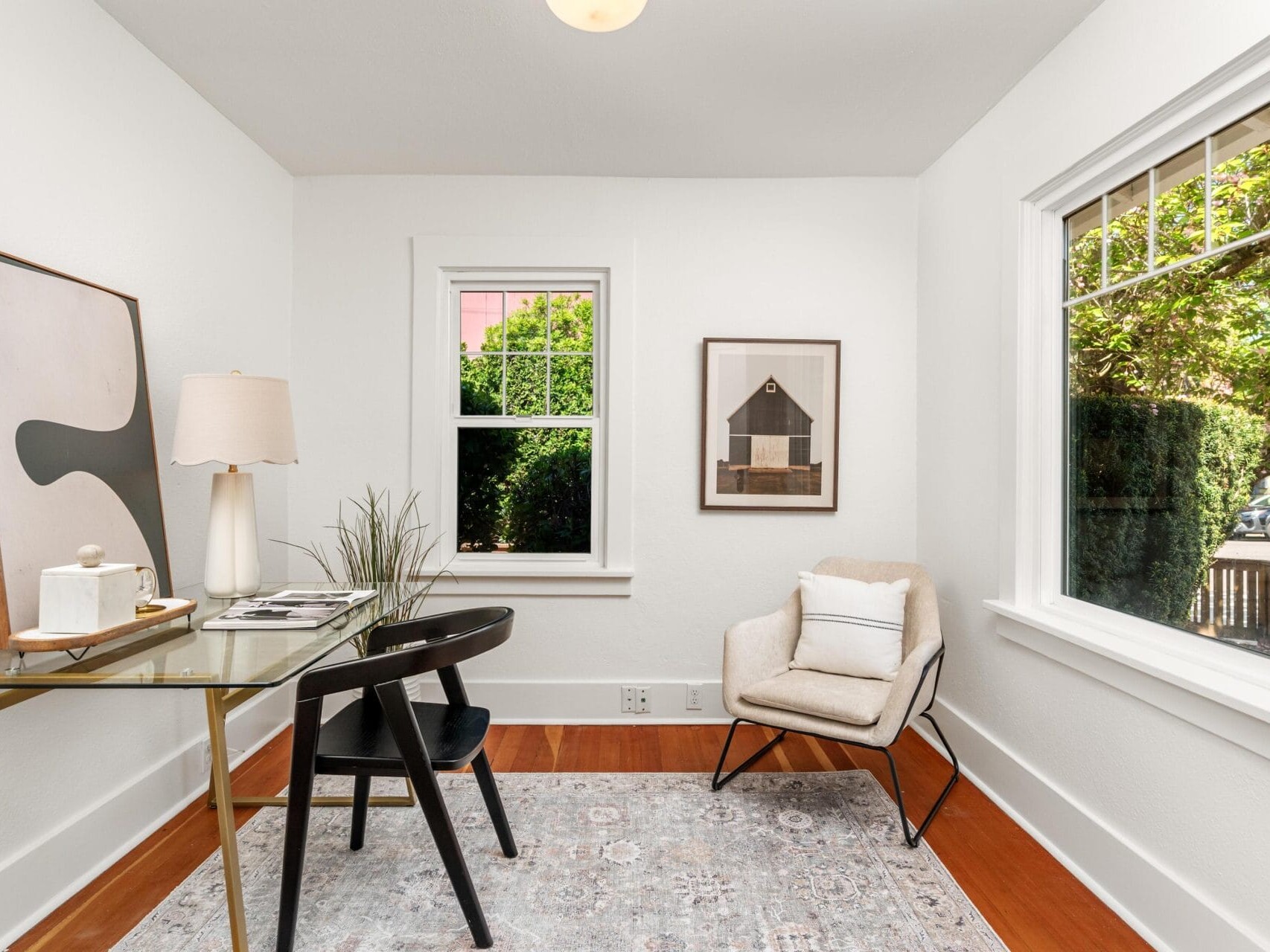 A bright home office with a glass desk, black chair, beige armchair, modern art on the walls, a lamp, and large windows letting in natural light with green trees visible outside.