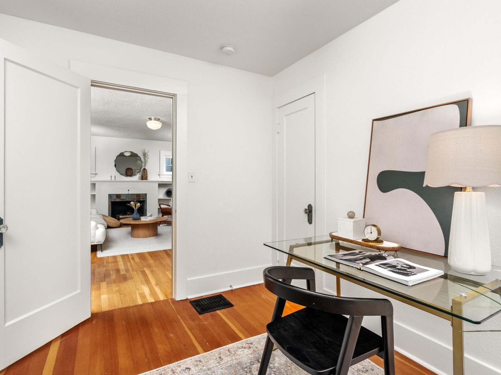 A small, modern home office with a glass desk, black chair, table lamp, and abstract artwork. The room has white walls, wood floors, and doors leading to a cozy living room with a fireplace.
