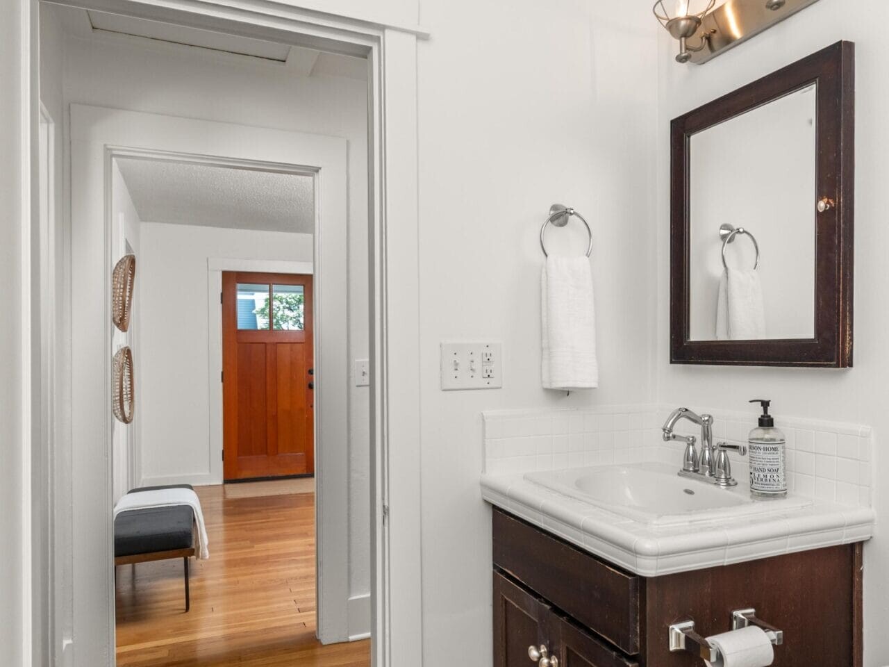 A small bathroom with a dark wood vanity, white sink, wall mirror, and two light fixtures. The view extends into a hallway with wood floors and a front door with windows.