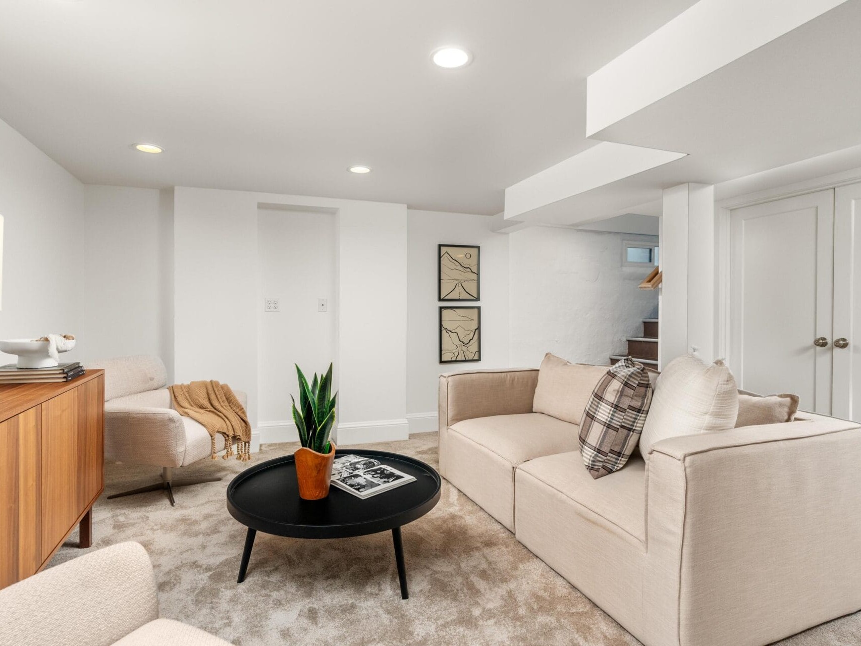A modern, cozy basement living room with beige sofas, a round black coffee table, a wooden sideboard, light carpet, abstract wall art, and a potted plant, lit by recessed ceiling lights.