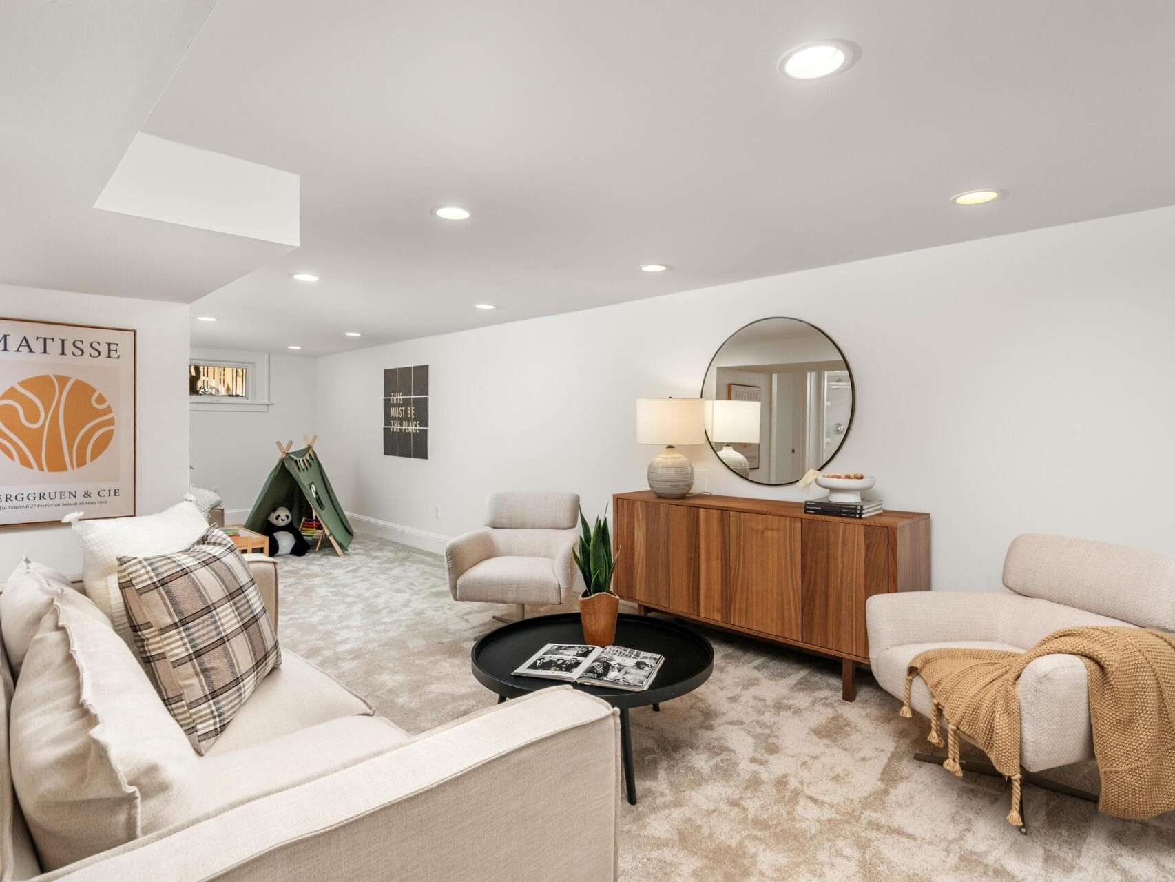A modern, cozy living room with beige carpet, white walls, a light sofa, armchairs, a round black coffee table, a wooden sideboard, a large round mirror, wall art, and warm lighting. A blanket and plant add a homey touch.