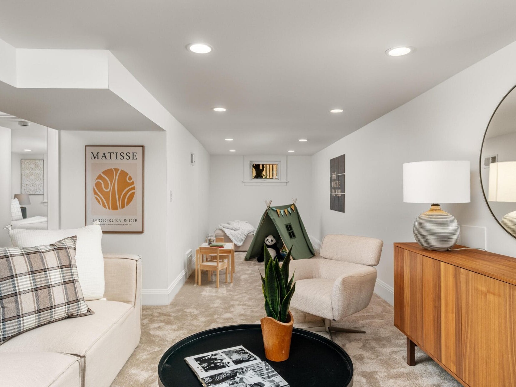 A modern, cozy living room with beige carpet, cream furniture, a round mirror above a wooden sideboard, a potted plant, a small play tent, kids table and chairs, and wall art featuring a Matisse print.