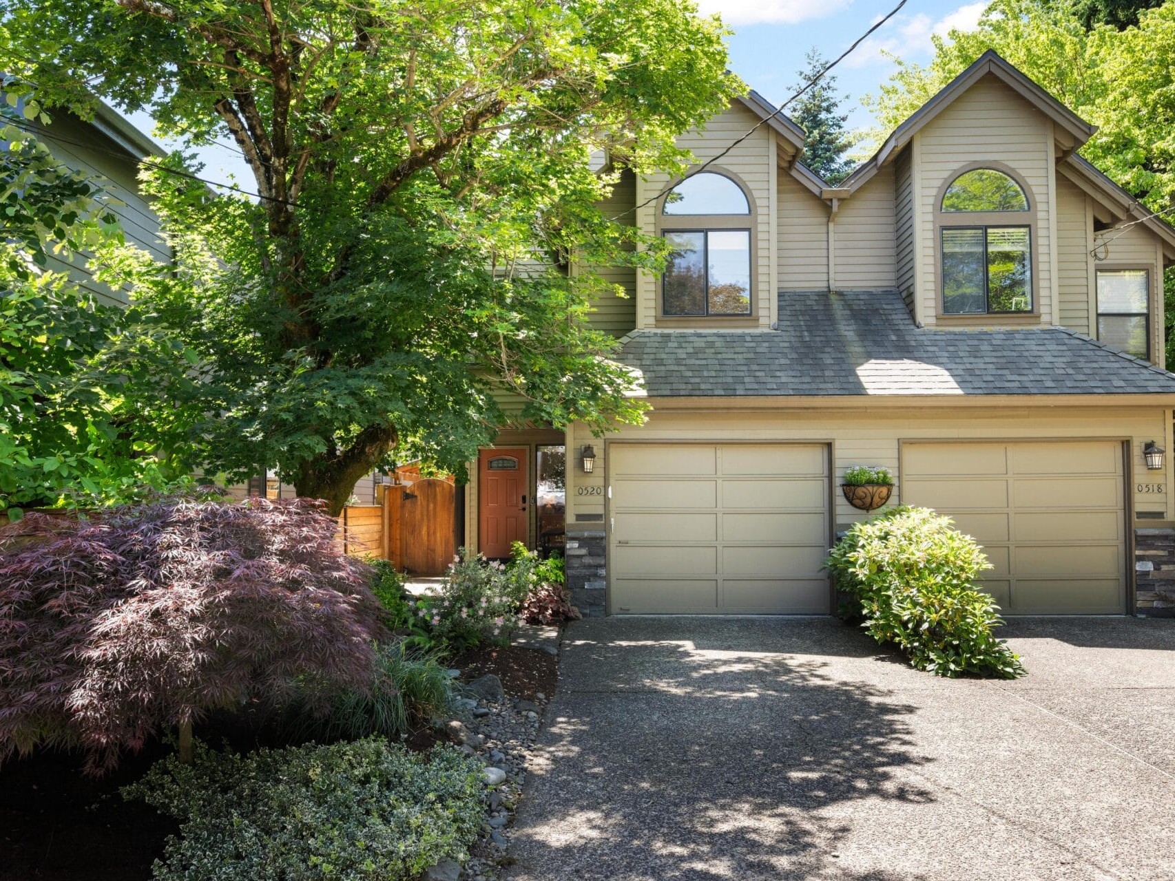 Two-story suburban house with light gray siding, double garage doors, arched windows, and a tree-shaded driveway surrounded by lush green landscaping and shrubs in the front yard.