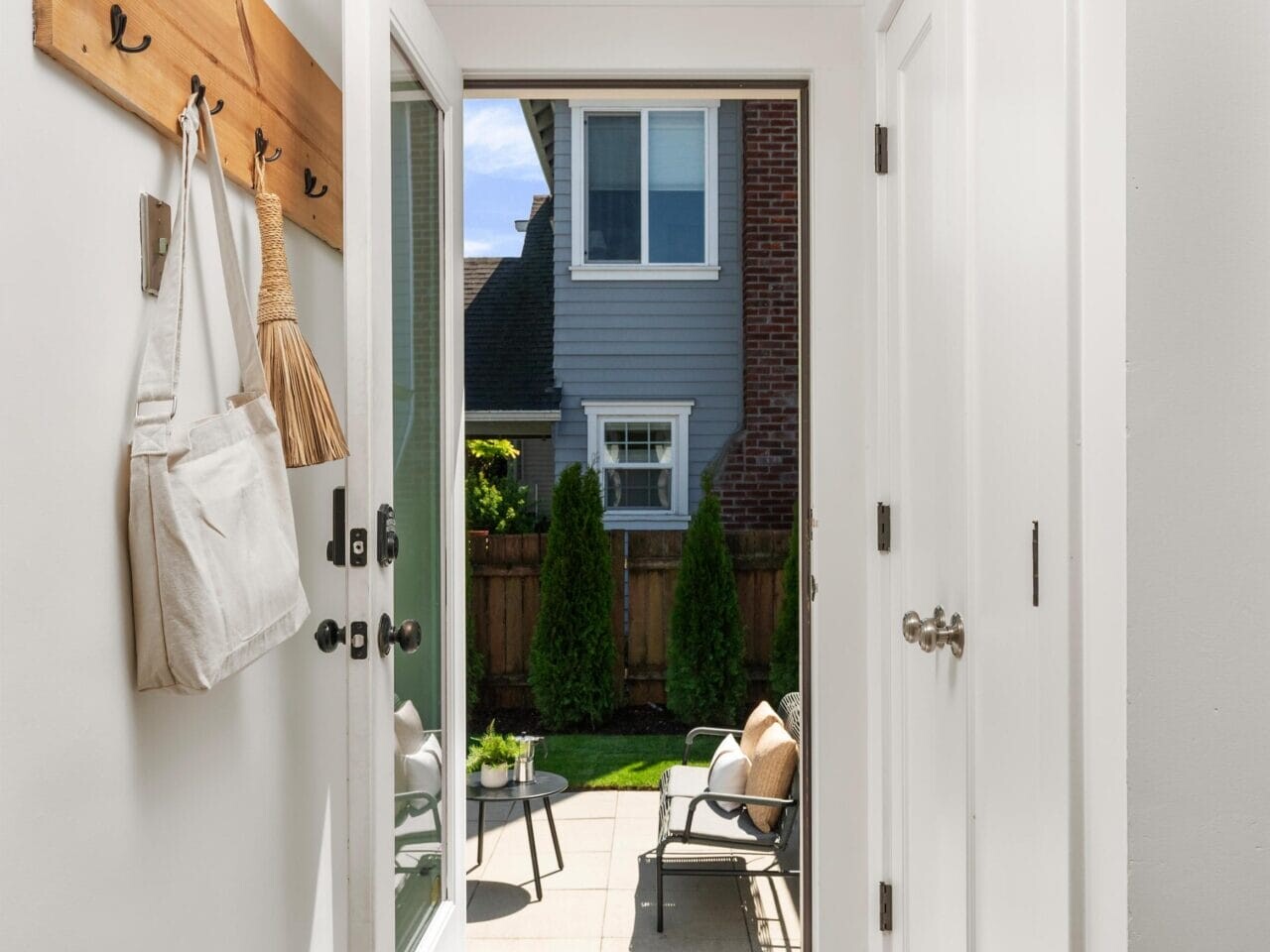 A bright entryway with white walls and doors, a wooden coat rack holding bags, and an open door leading to a sunny patio with outdoor chairs, a table, and green shrubs against a fenced yard.