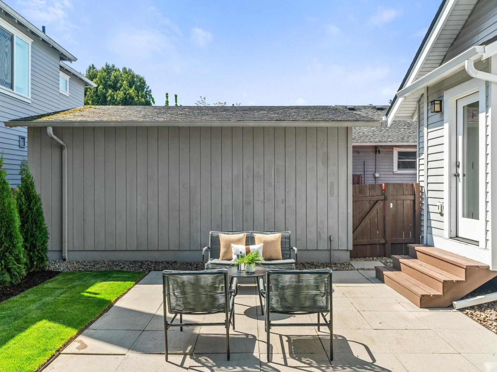 A small backyard patio with gray pavers, a glass table and two chairs, beige cushions, and a few potted plants. The area is bordered by grass, a wooden fence, and a gray building. Steps lead up to a white house.