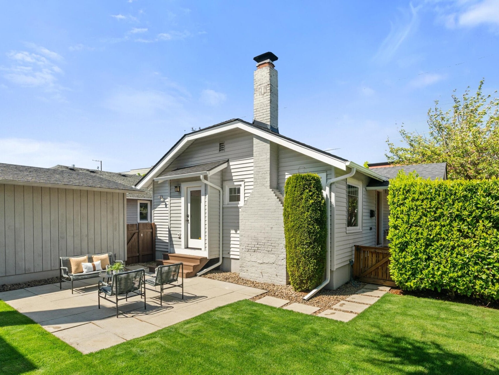 A small, light-gray house with a white brick chimney, a neatly trimmed hedge, and a green lawn. There is a patio area with outdoor seating and a table on a sunny day.