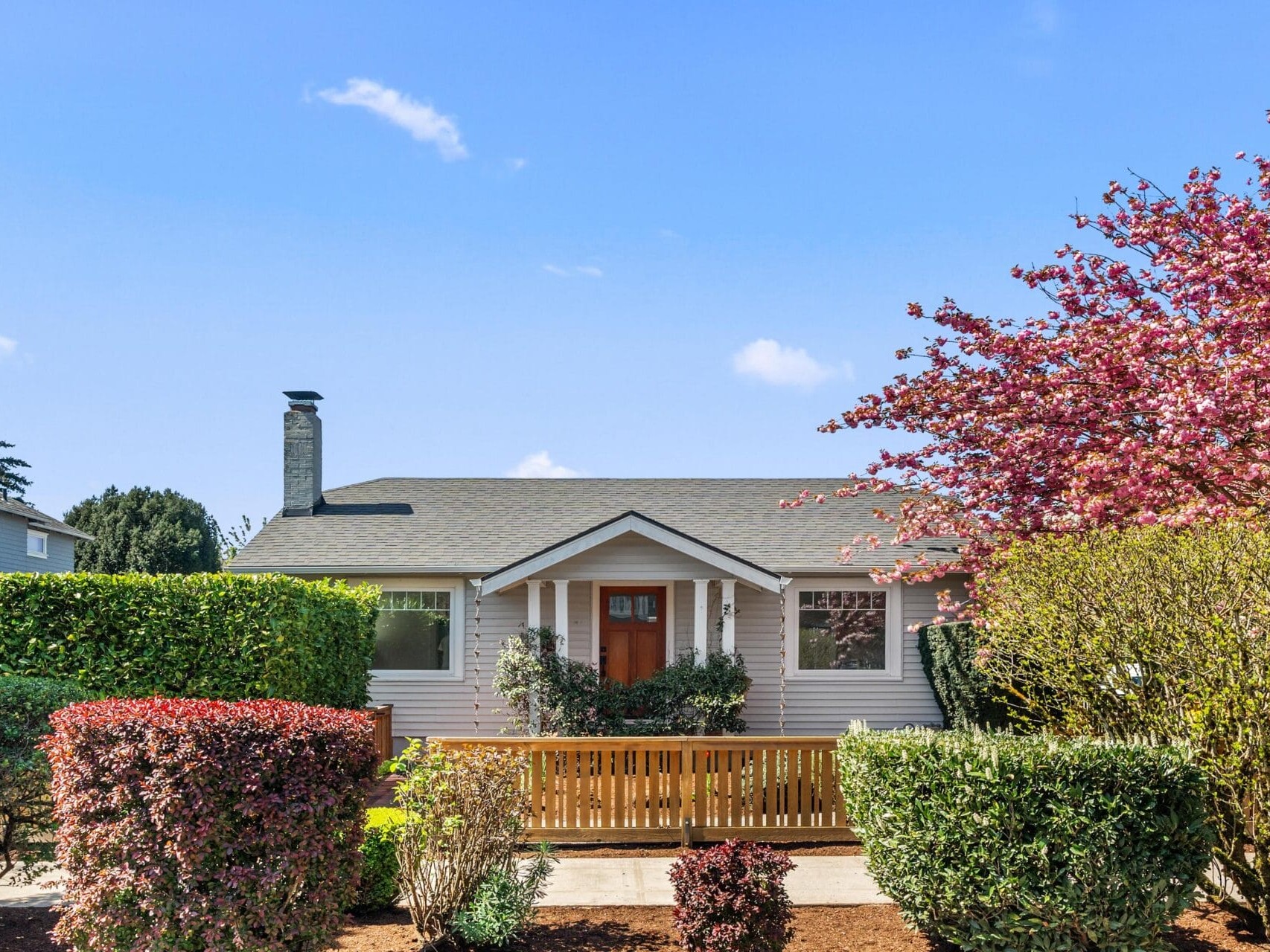 Single-story house with light-colored siding, a gray shingle roof, and a small front porch. The front yard features neatly trimmed bushes and a blooming tree under a clear blue sky.