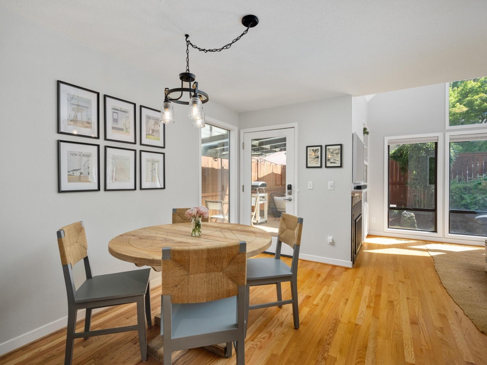 A dining area with a round wooden table, four chairs, wall art, and a modern black light fixture. Large windows and a glass door let in natural light, highlighting the hardwood floor and leading to a patio and a fenced yard.